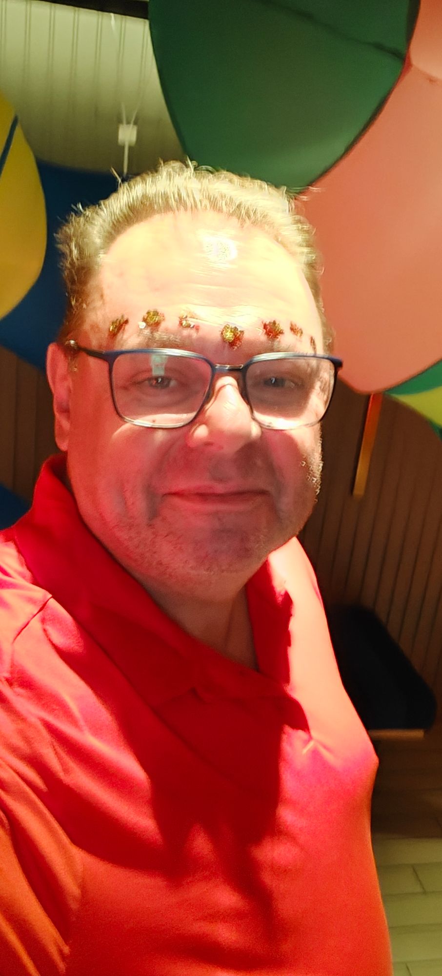 Man in red shirt and glasses smiling at camera with colorful balloons visible behind him in bright indoor setting