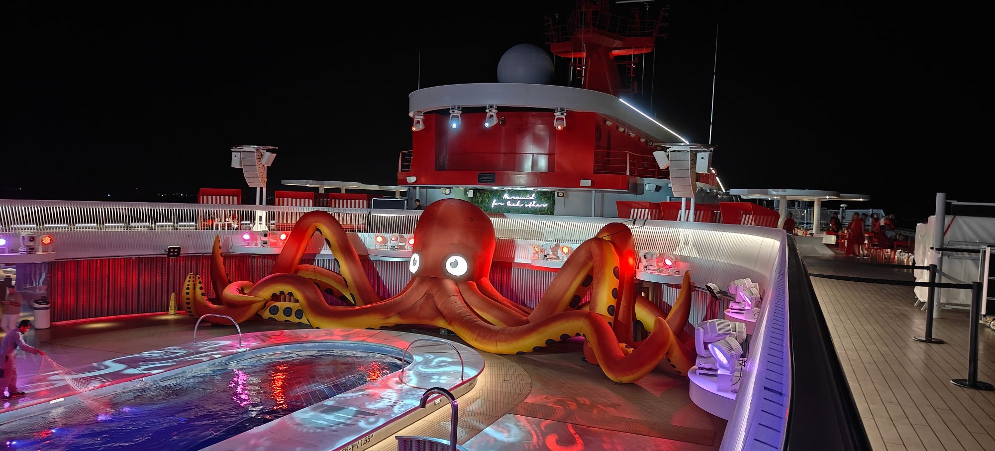 Nighttime deck view of large red octopus water slide sculpture illuminated in purple and red lighting near ship superstructure