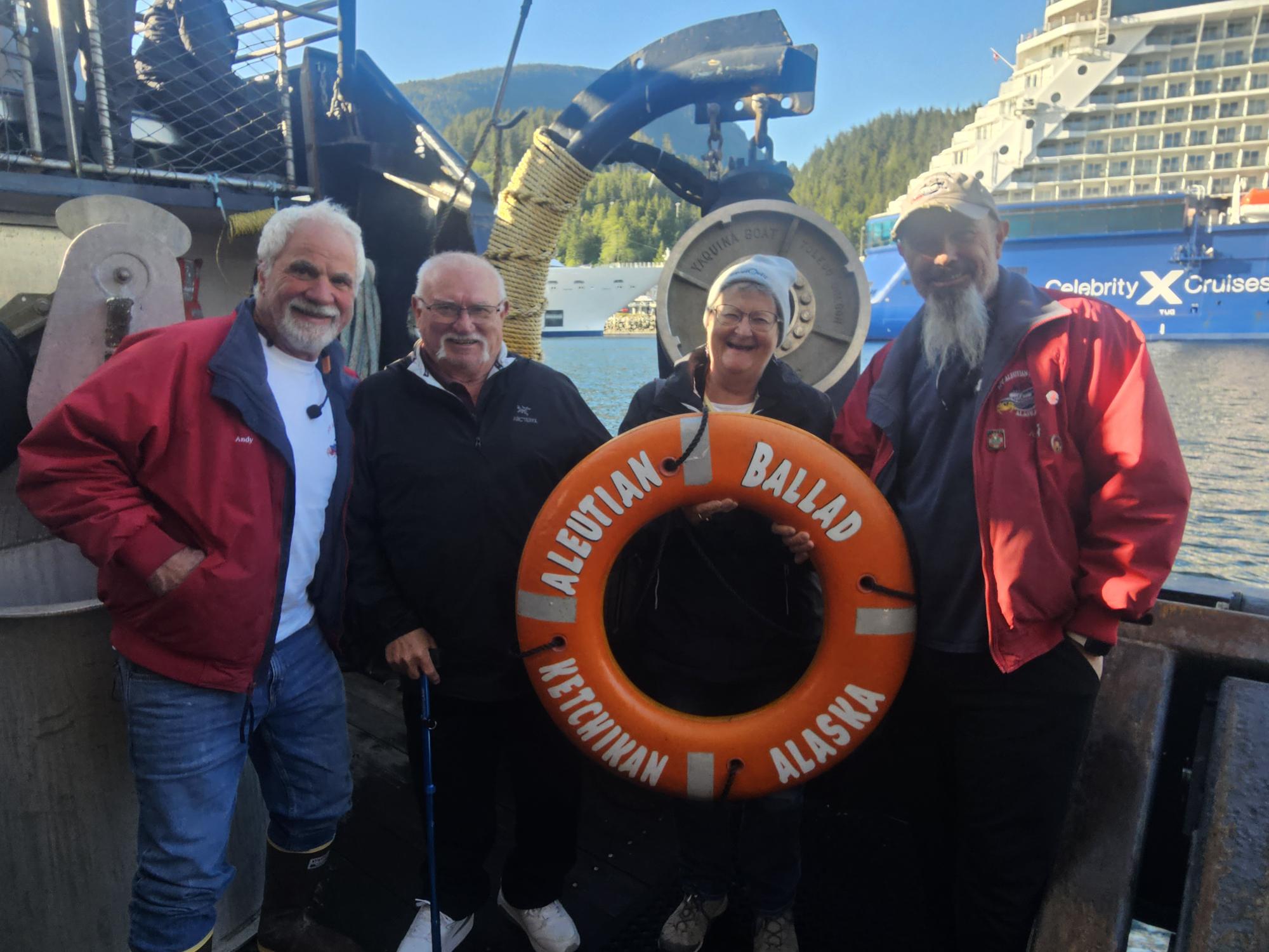 Four people in red jackets holding a life ring labeled 'Aleutian Ballad' aboard a ship in Alaska