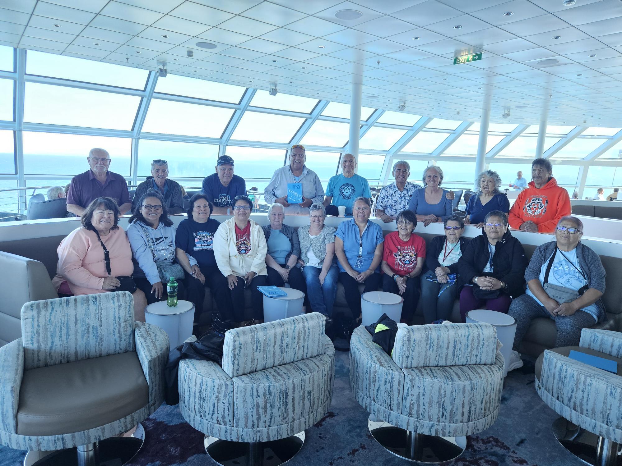 Group of cruise passengers gathered in a bright observation lounge with ocean views visible through large windows