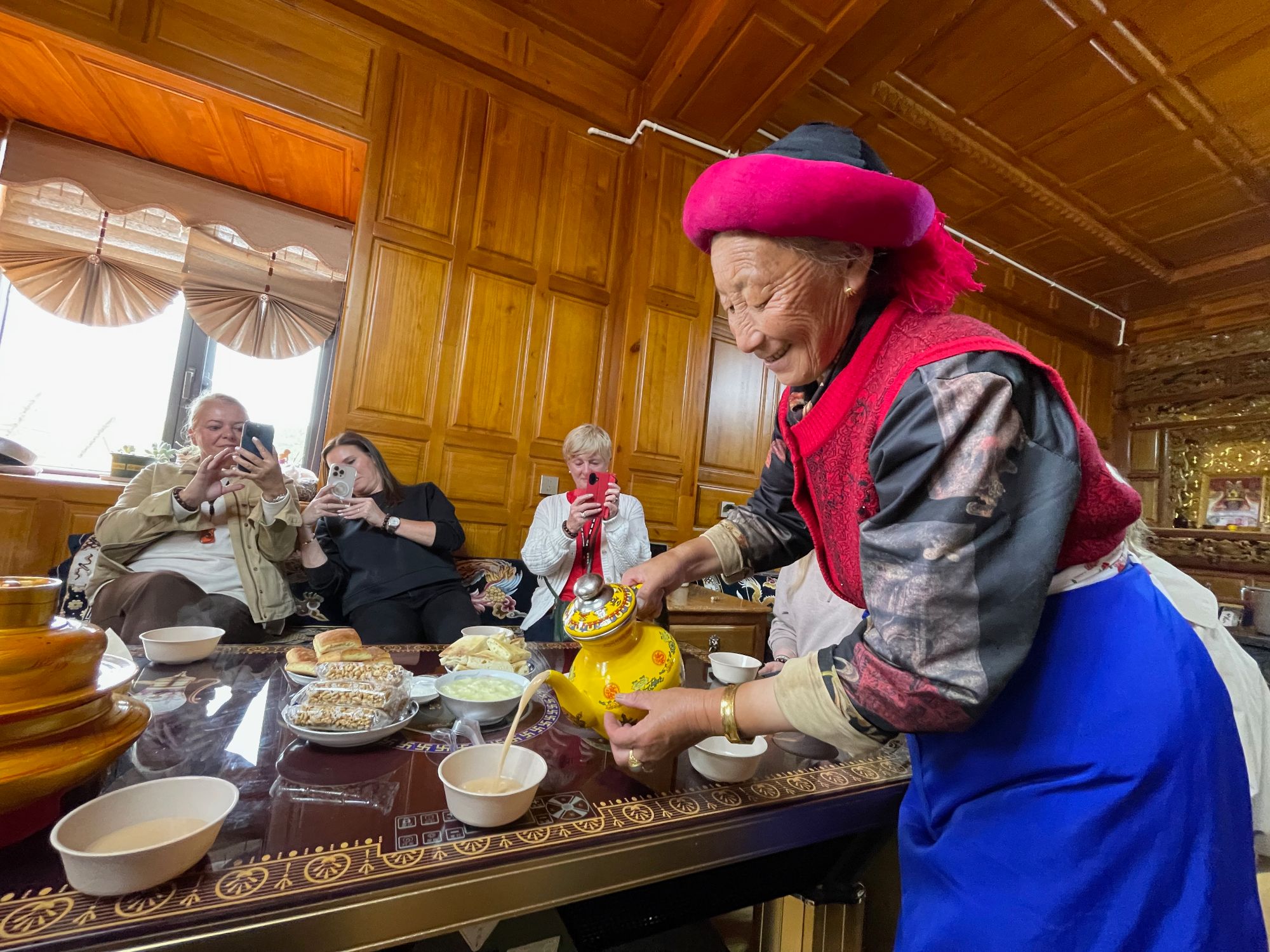 Elderly woman in traditional Chinese clothing serving food to guests in a wooden-paneled dining room
