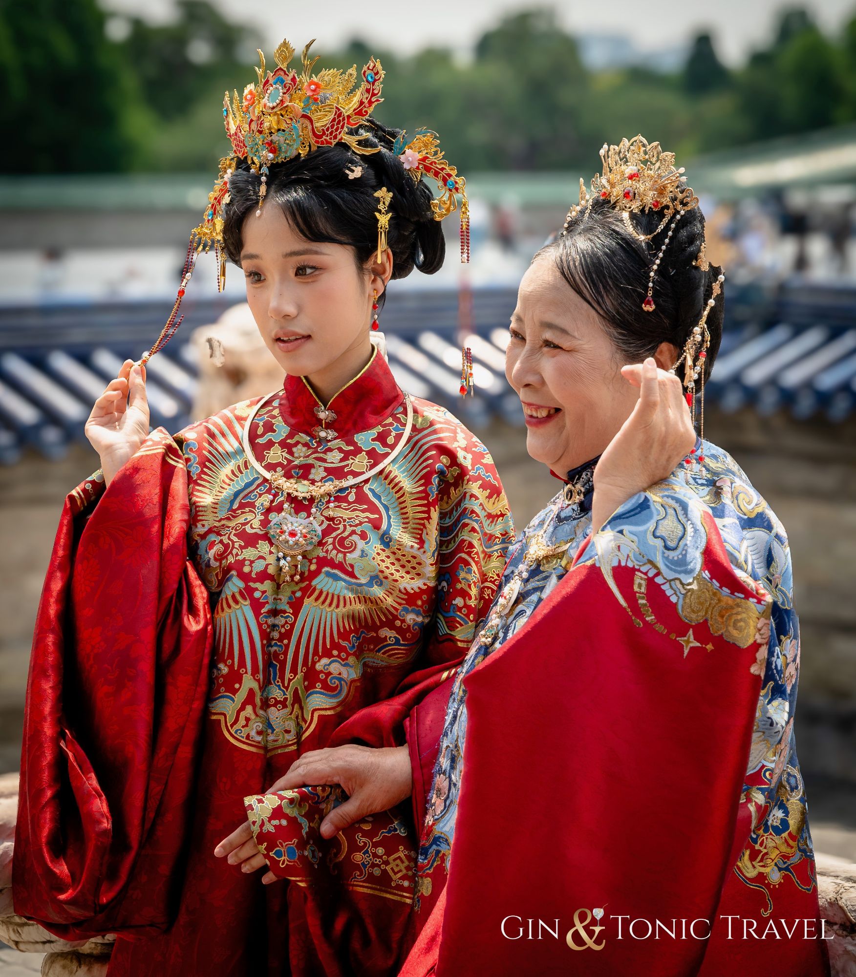 Two women wearing elaborate red silk robes with gold embroidered phoenix designs and ornate headdresses by a waterfront