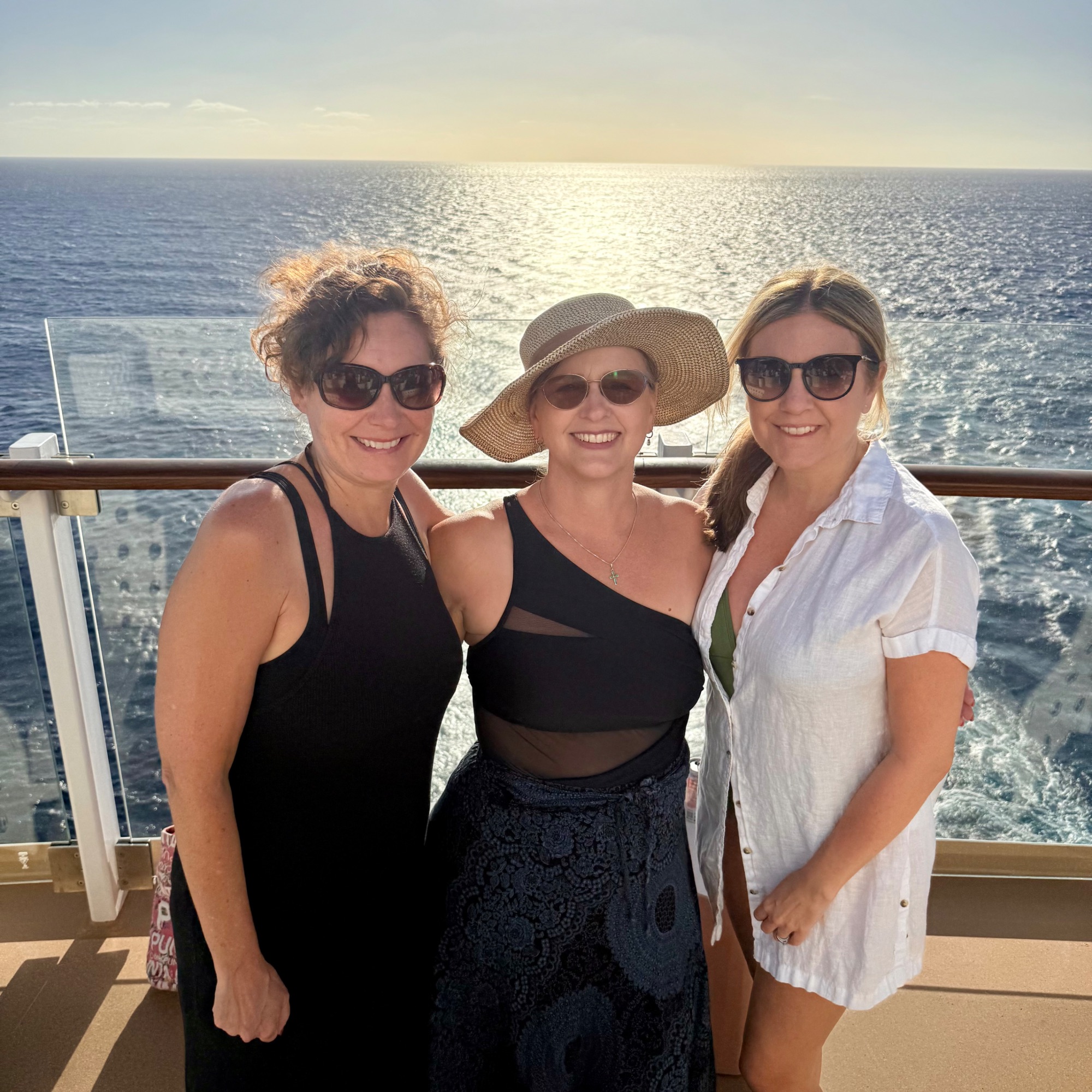 Three women in black swimwear stand at a ship railing overlooking calm ocean waters