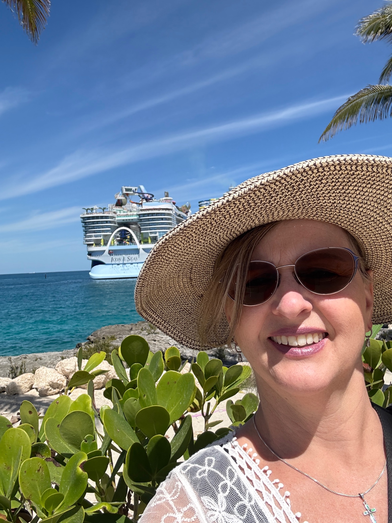 Woman in wide-brimmed straw hat and sunglasses smiles in front of a large cruise ship docked at a tropical port