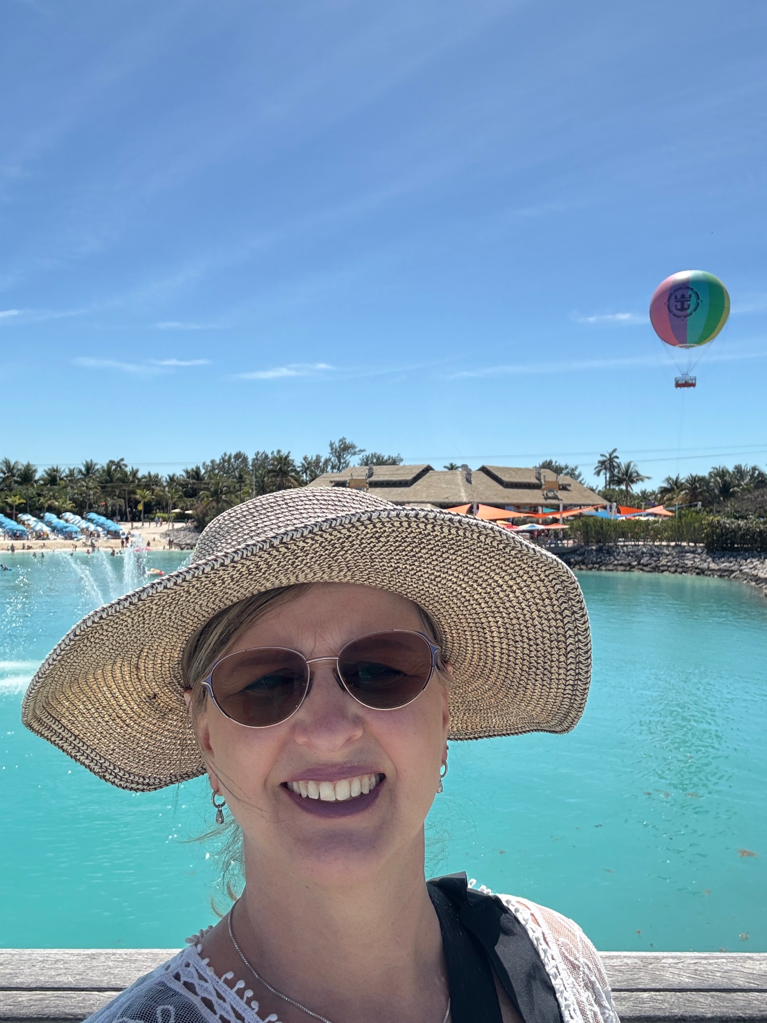 Woman in straw hat poses by a turquoise lagoon with a hot air balloon visible in the sky