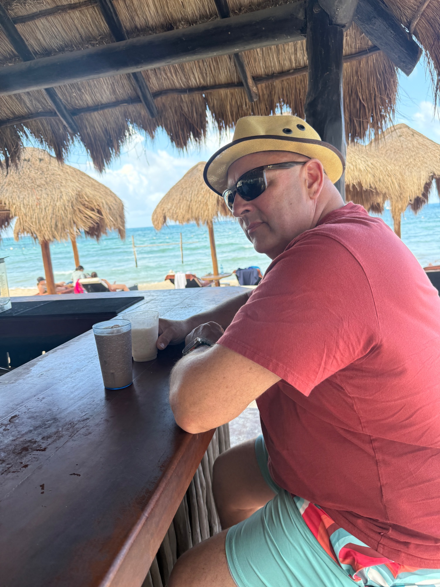 Man in yellow hat and sunglasses relaxing at a beach bar with thatched umbrellas and ocean view