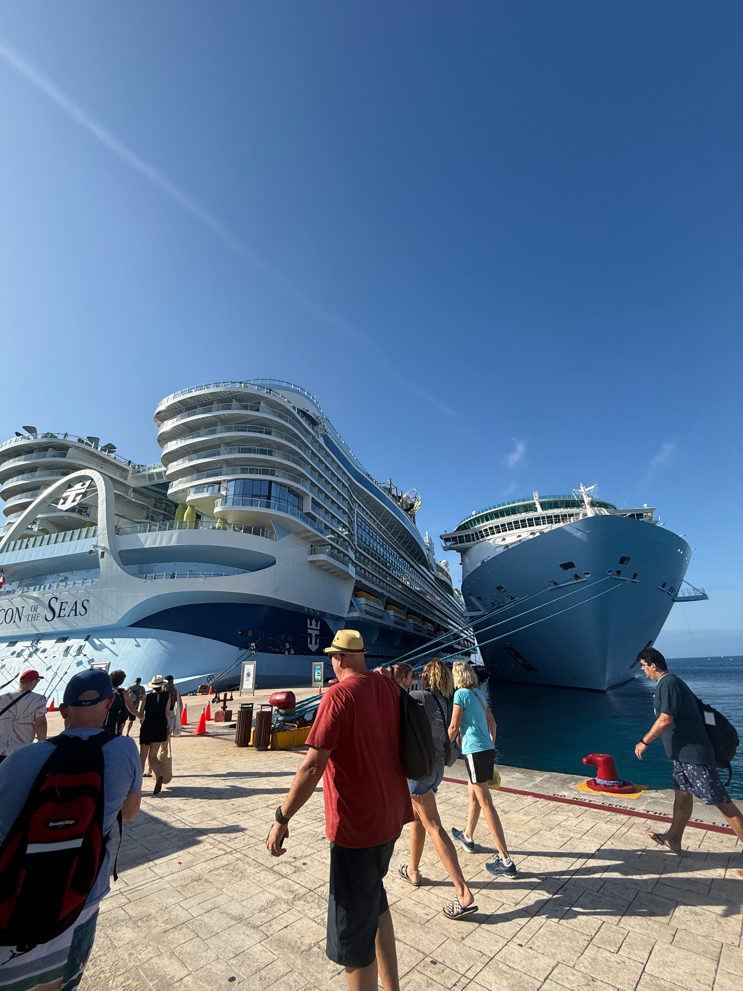 Two large cruise ships docked side by side at a Caribbean port with passengers walking nearby