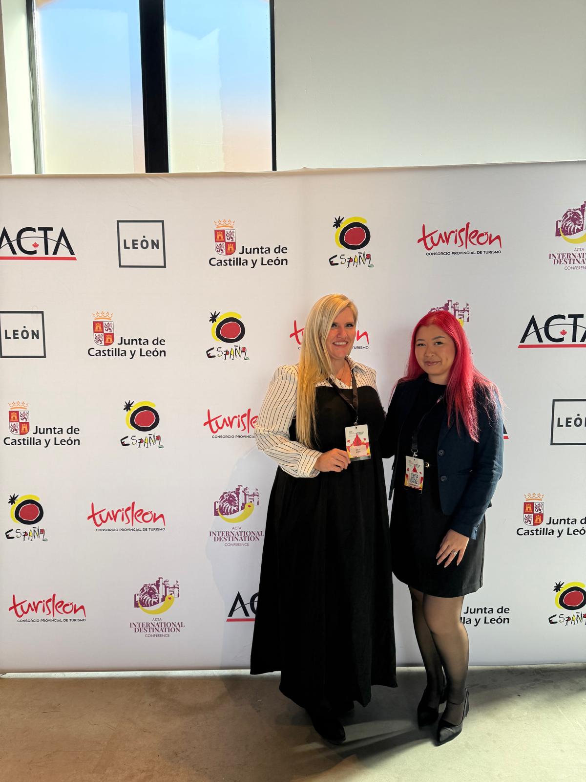 Two women wearing event badges pose together in front of a sponsor backdrop at an indoor tourism conference