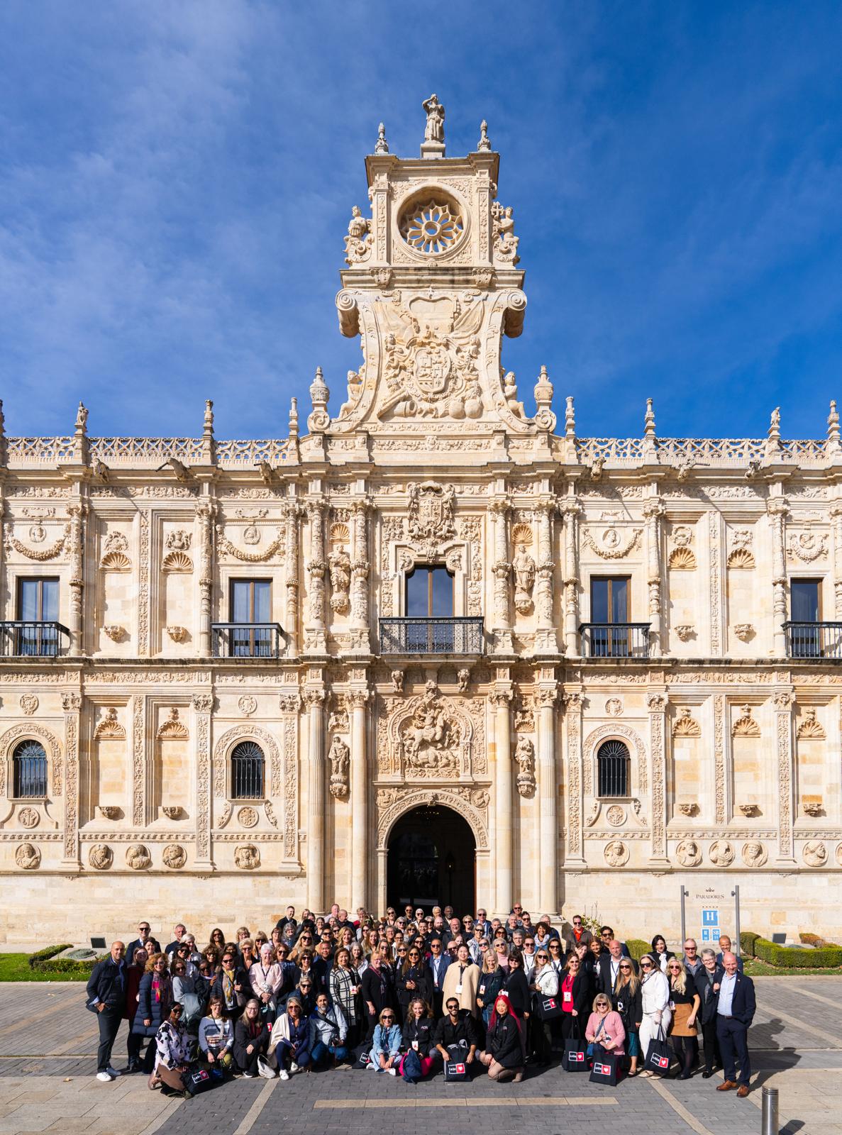 Large group of tourists with event badges gathered in front of an ornate Renaissance palace with a clock tower in León, Spain