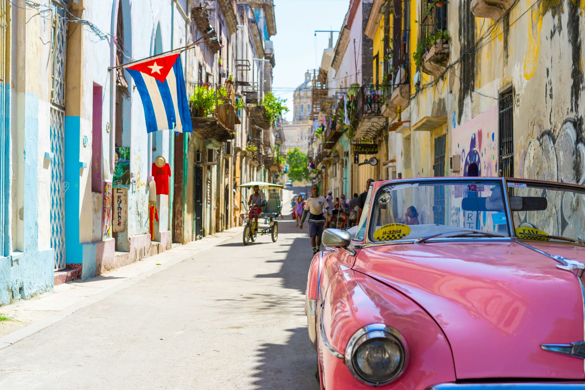 A pink vintage American car parked on a narrow Havana street beneath a Cuban flag hanging between colorful buildings