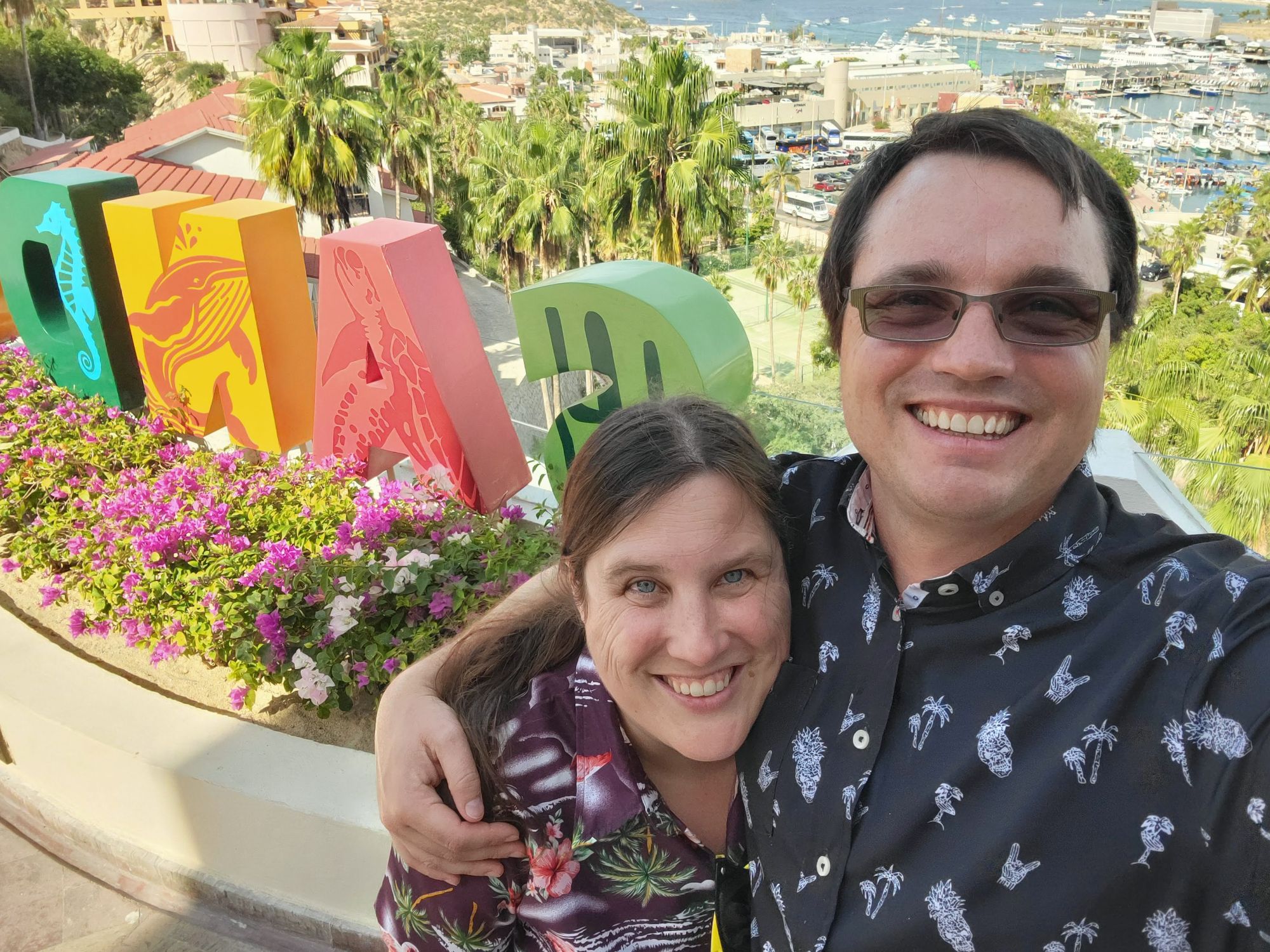 Kayleen Hamelin and a companion smile at a colorful art installation overlooking a marina and palm trees in Cabo