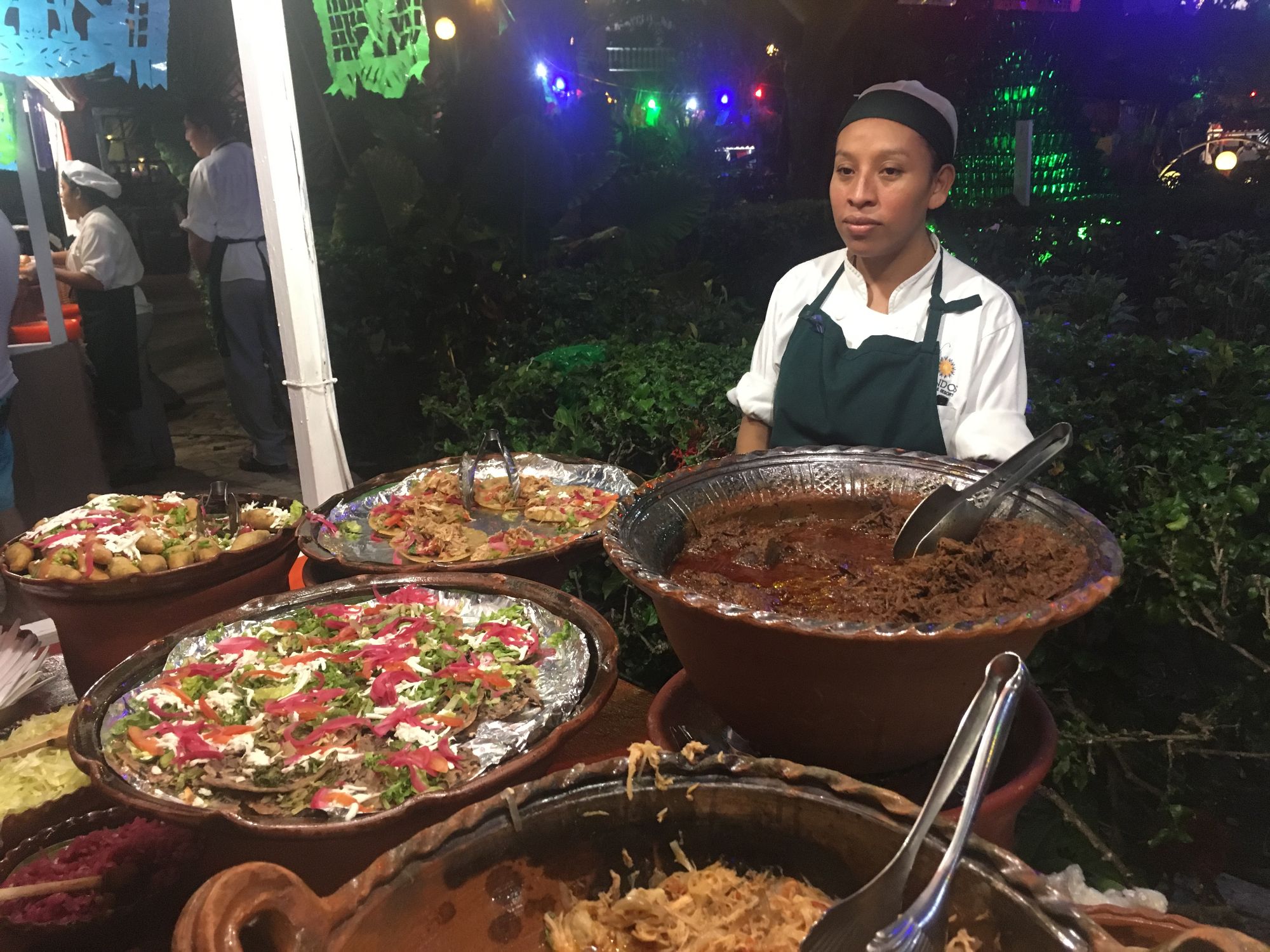 Server in apron standing behind traditional Mexican dishes including mole and colorful prepared foods at evening event