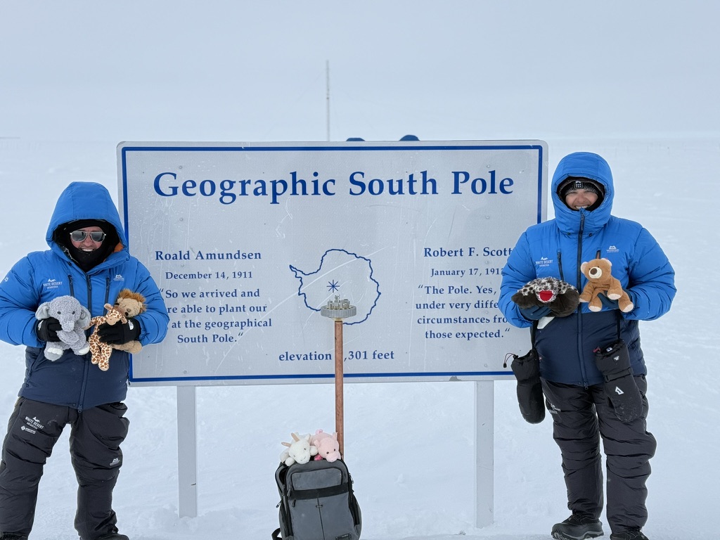 Two people in blue jackets hold stuffed animals at Geographic South Pole marker sign