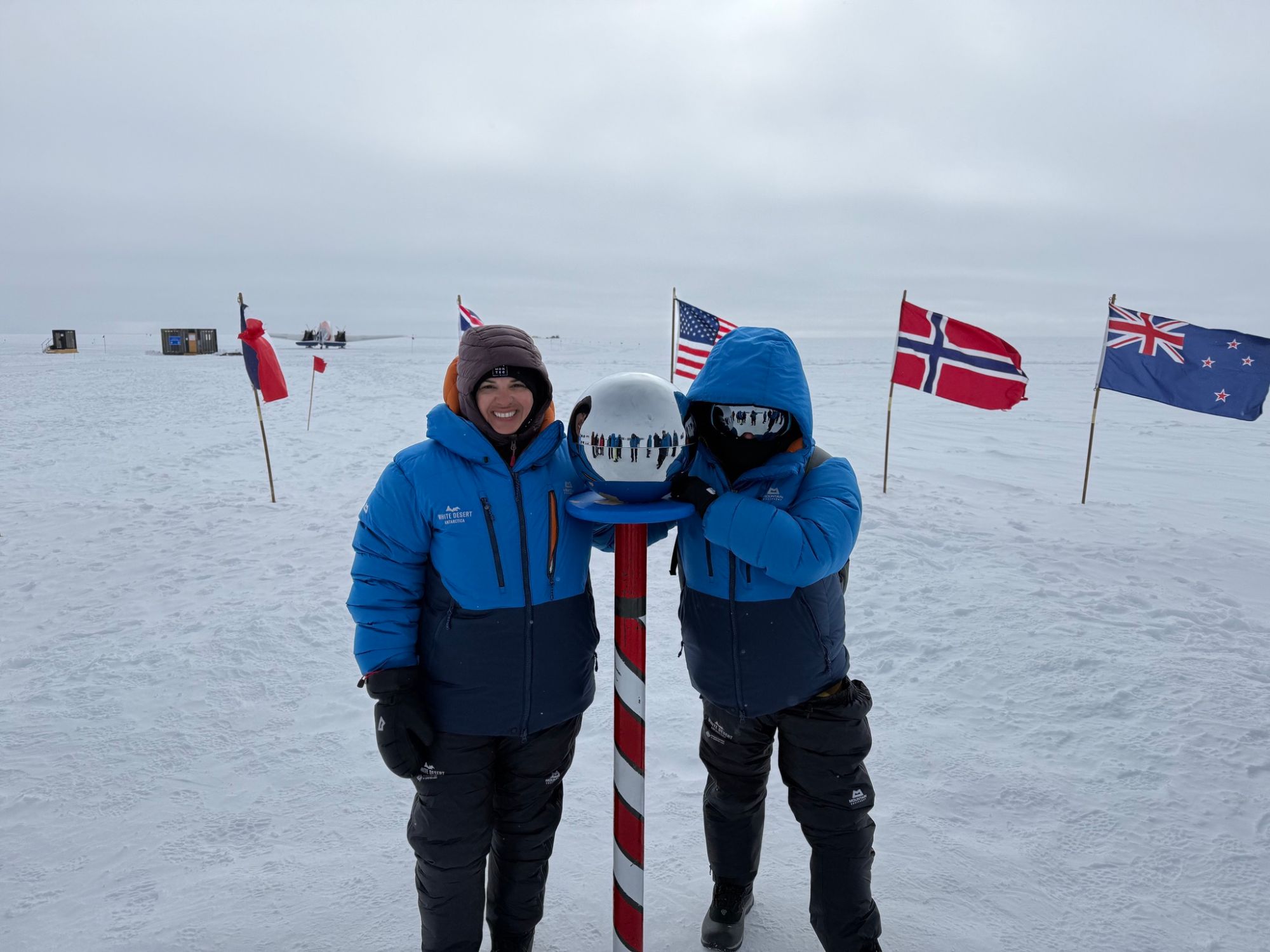 Two people in blue jackets pose with sphere and international flags at South Pole
