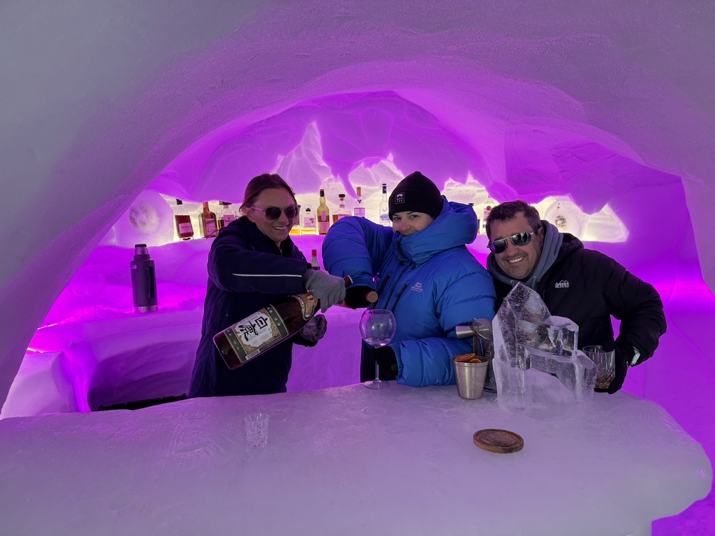 Three people inside ice cave with magenta neon lighting, holding drinks at bar