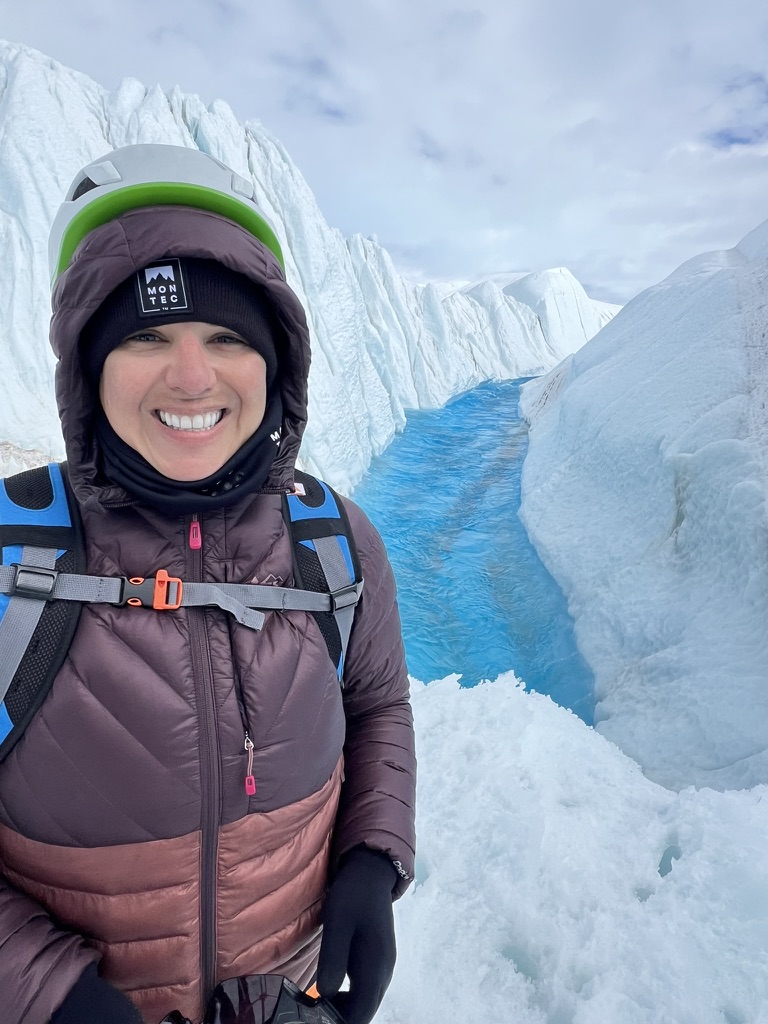 Diania Pimenta smiles between tall blue-ice walls in Antarctica wearing climbing gear