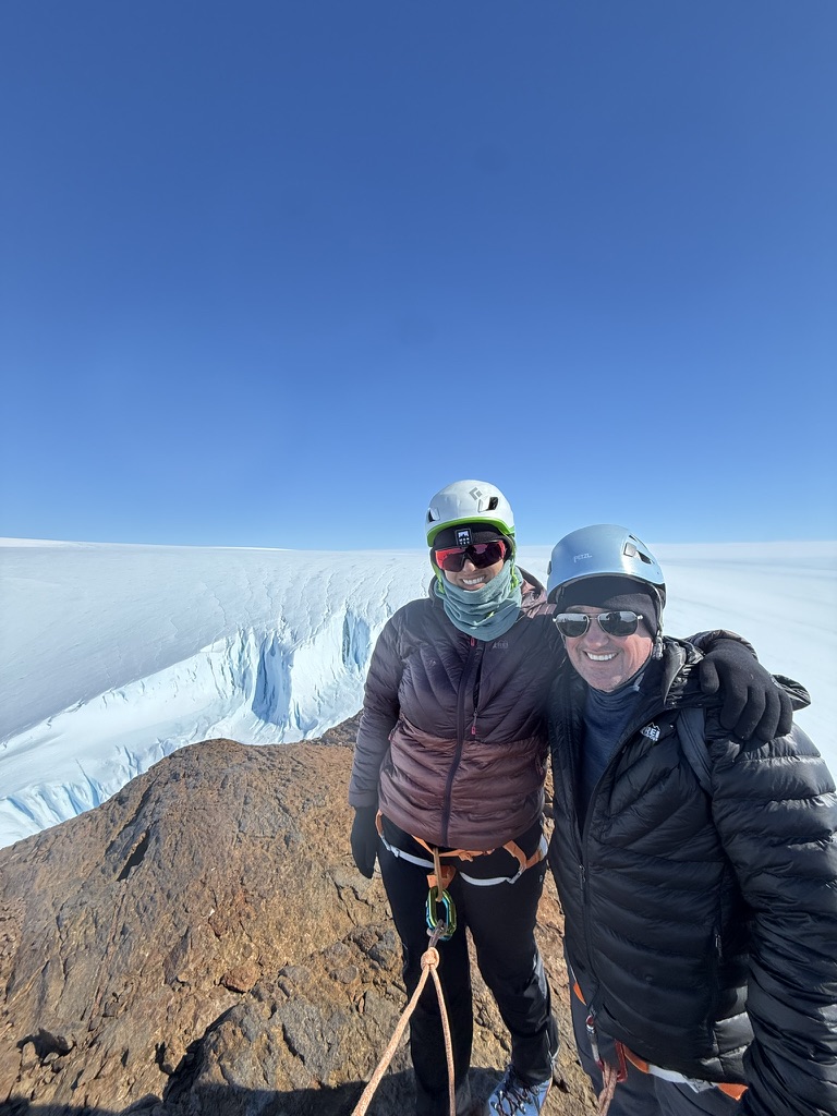 Two climbers in helmets pose together on rocky outcrop with glacier and ice wall behind