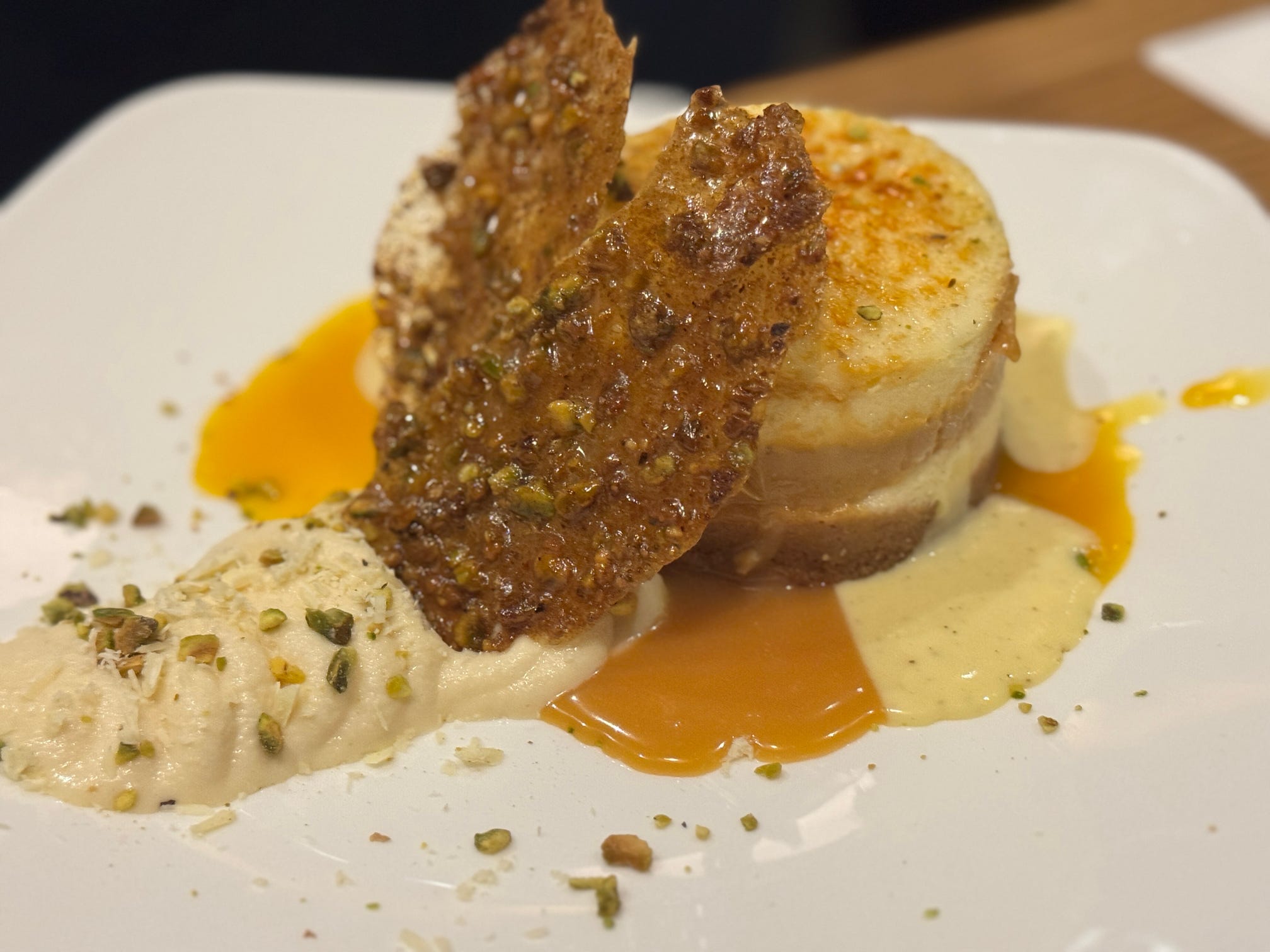 An elegant plated dessert featuring brûléed cream, pistachio brittle, and caramel sauce on a white plate.