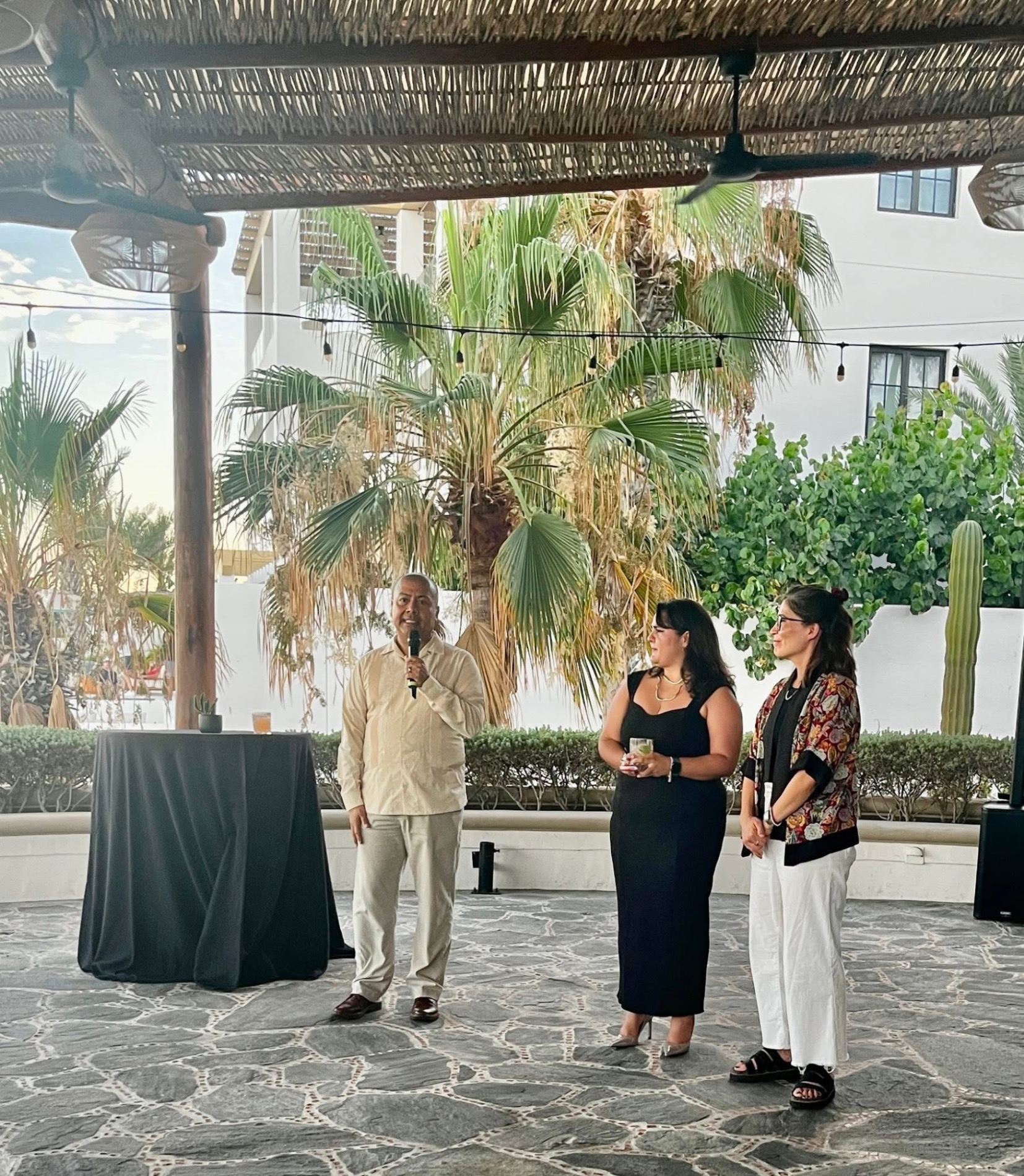 Man speaking at podium under woven shade structure with palm trees and white buildings behind