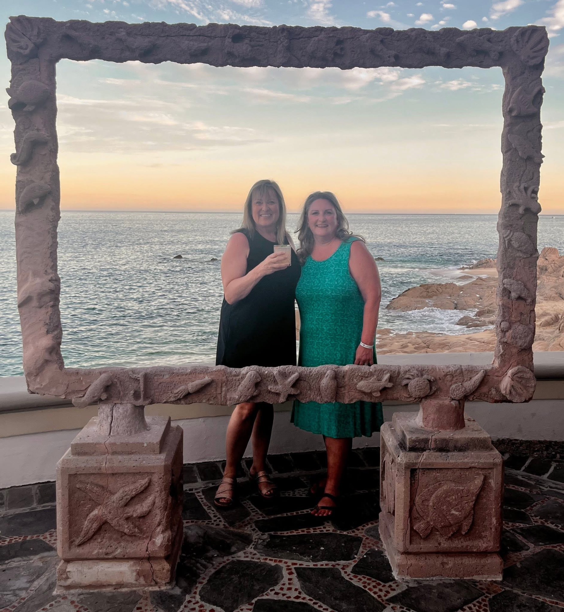 Two women posing in ornate stone frame with ocean and sunset in background