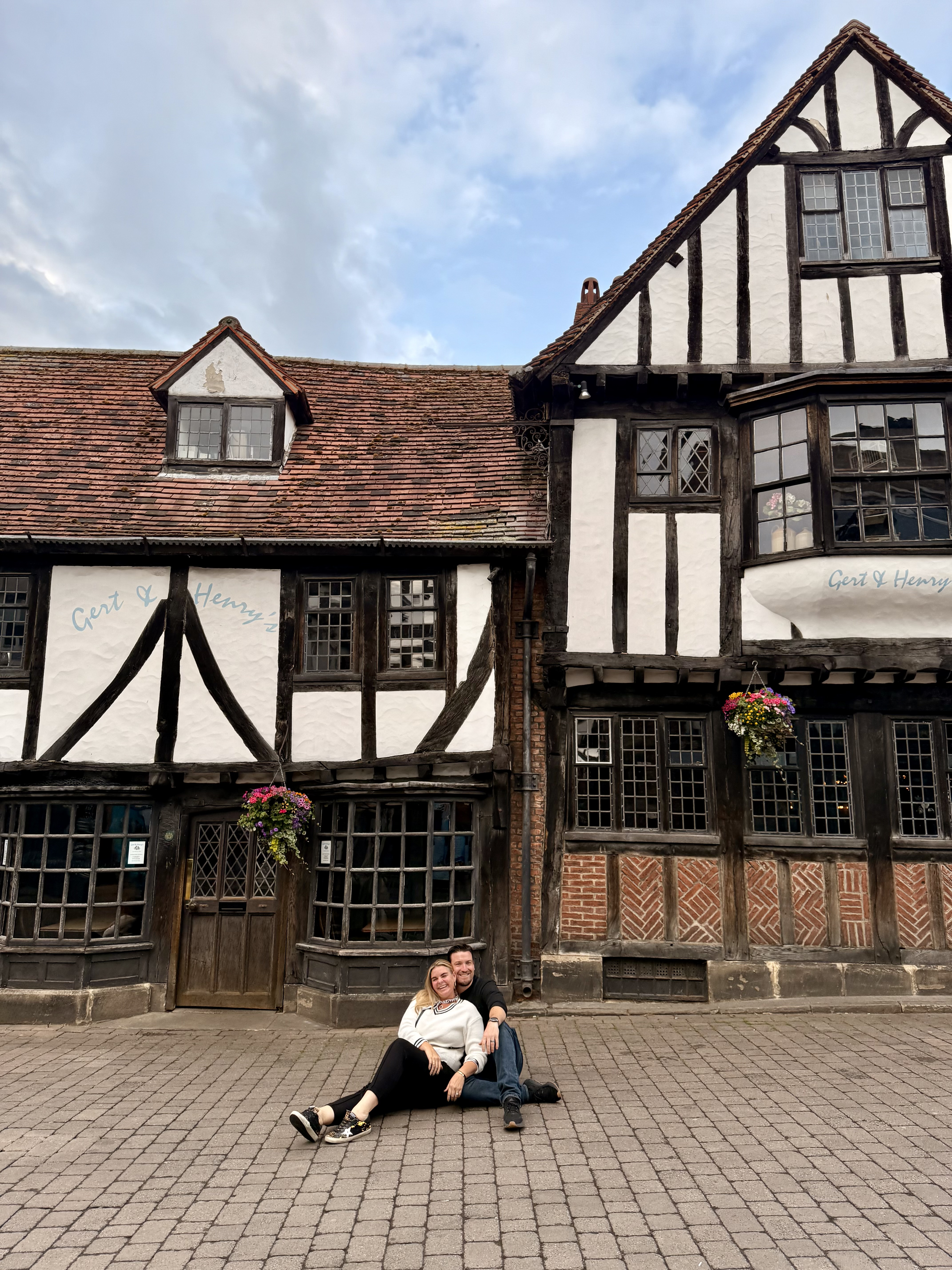Half-timbered Tudor building with white and dark wood framing, couple sitting on pavement in front