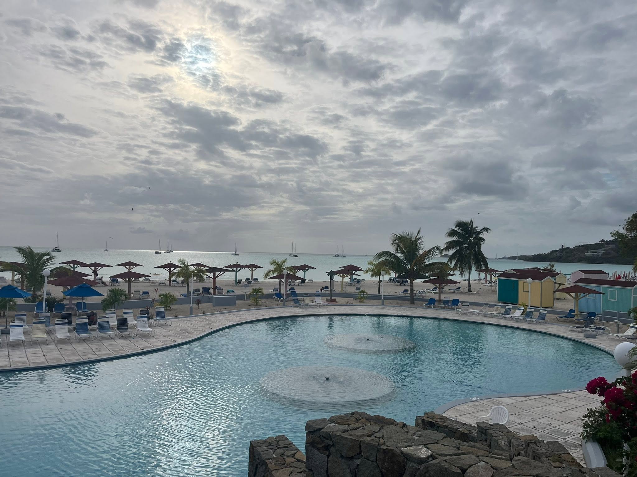 Resort pool with curved edges and central gazebo overlooking Antigua's beach, palm trees, and moored sailboats under cloudy skies