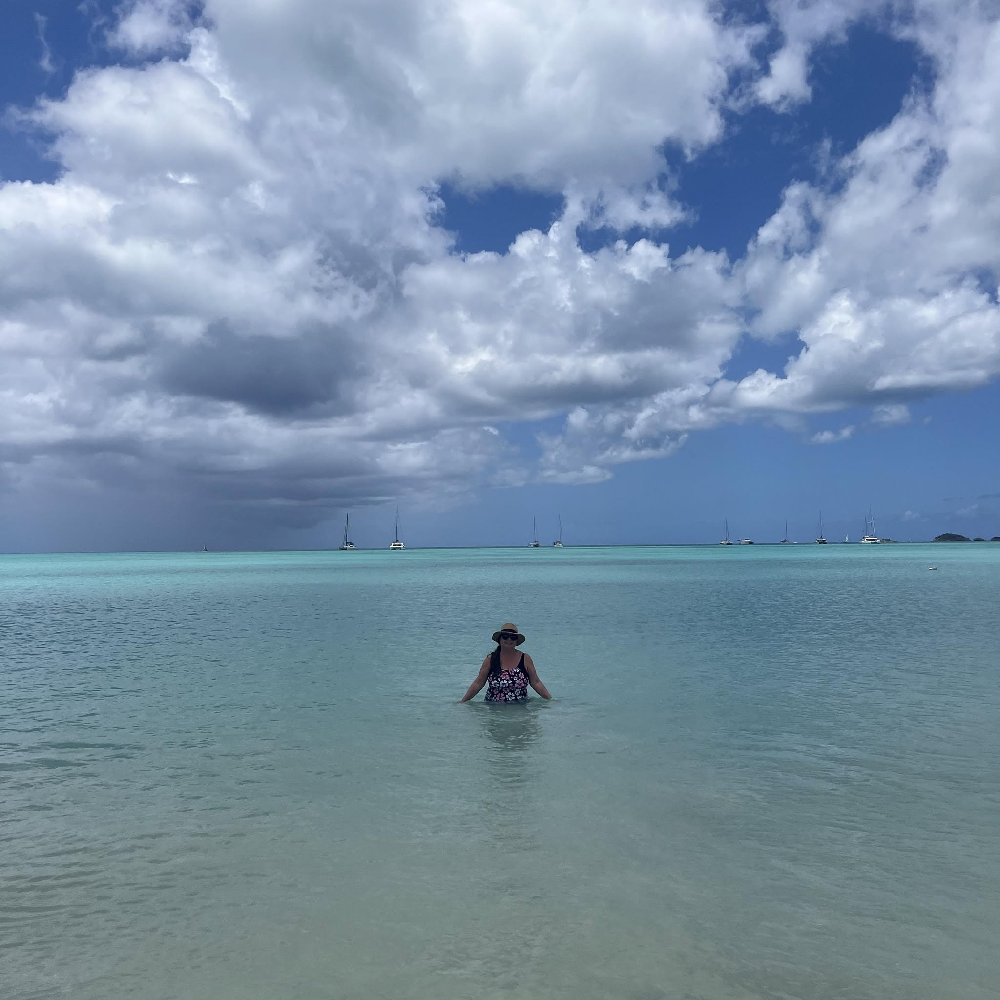 Woman wading in crystal-clear turquoise water off Antigua's beach with sailboats visible on the horizon and white clouds above