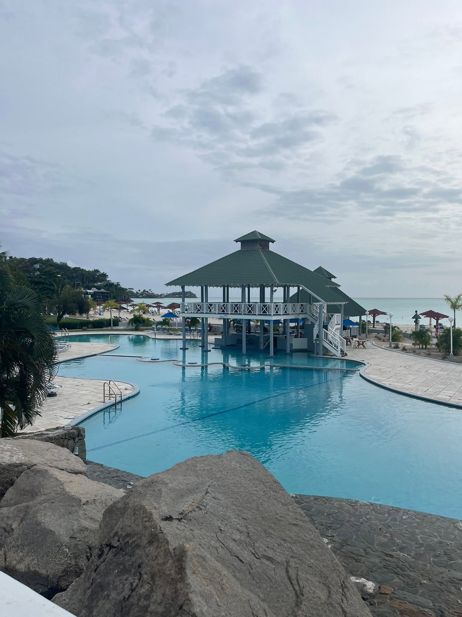 Resort pool complex with central octagonal gazebo bar, surrounding lounge chairs, and Antigua's beach and ocean visible beyond