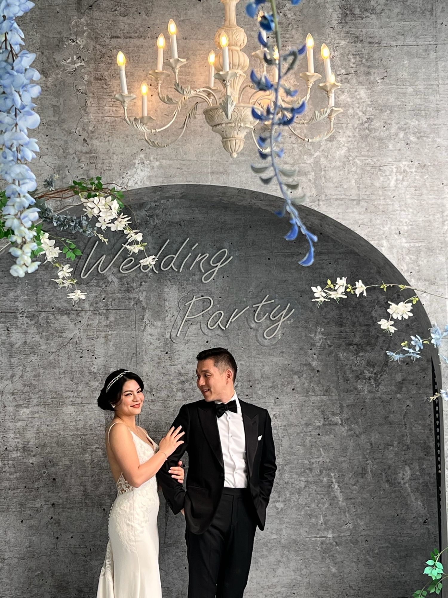 Kelly and her partner pose at the same wedding party setup, smiling while standing beneath an ornate cream-colored chandelier