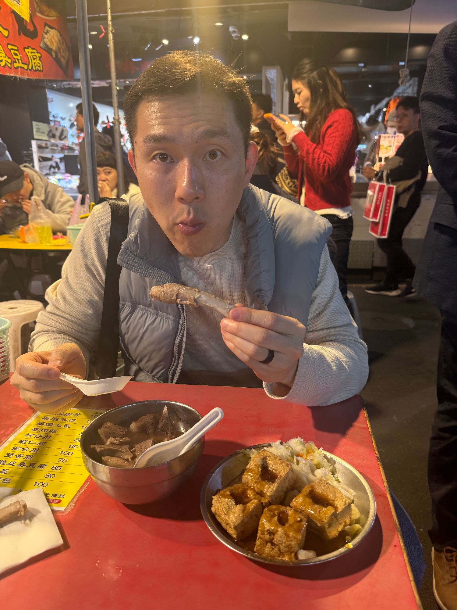 A man makes an exaggerated surprised expression while eating at a night market food stall with red signage visible