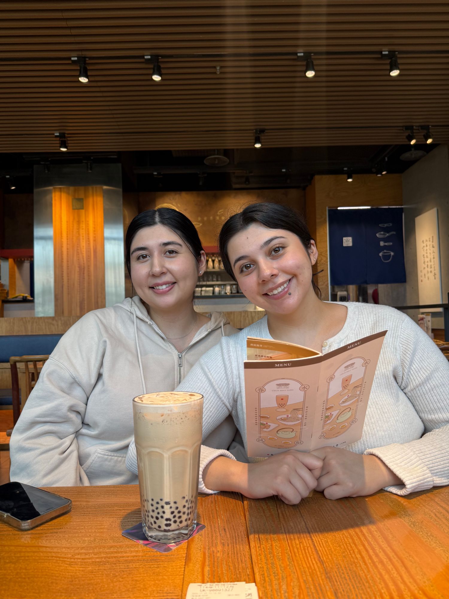 Two women sit at a restaurant counter holding a menu and a bubble tea drink, both smiling at the camera