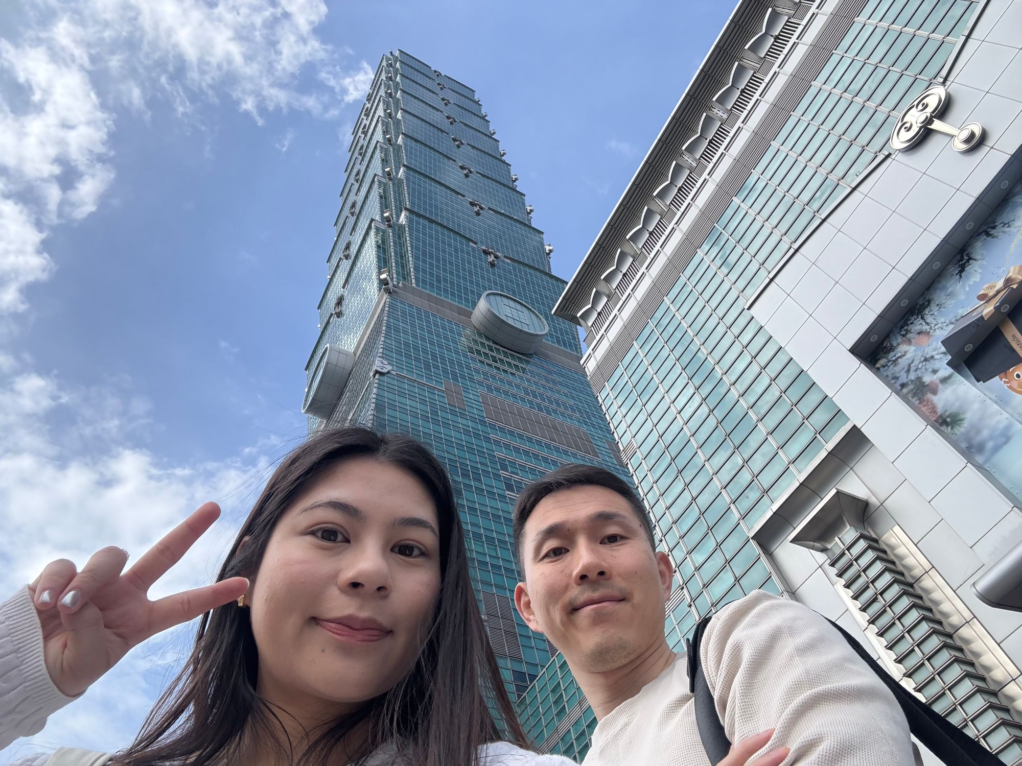 Kelly and a man stand together on a city street with a tall modern skyscraper visible behind them against a blue sky