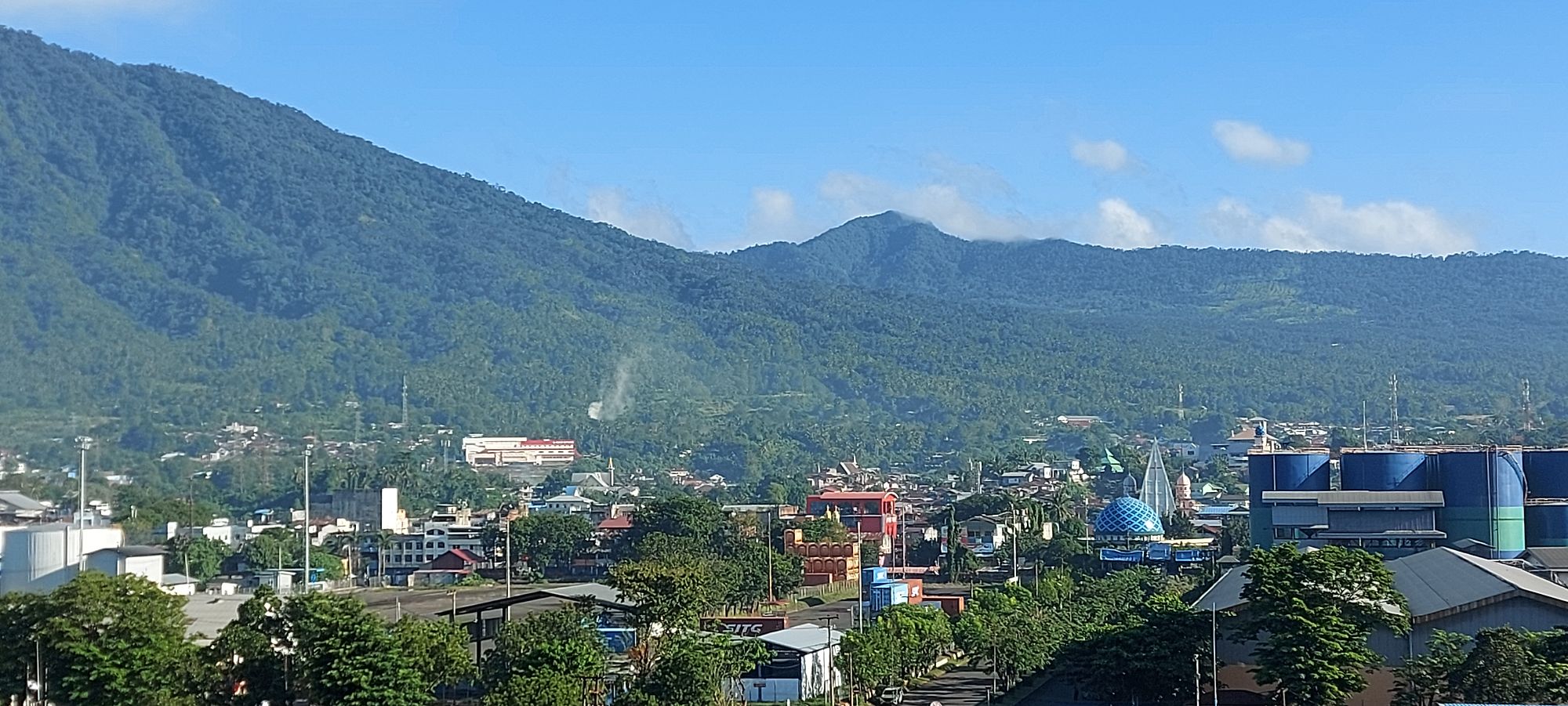 Densely forested mountains rising above Bitung city with scattered buildings and palm trees in the valley