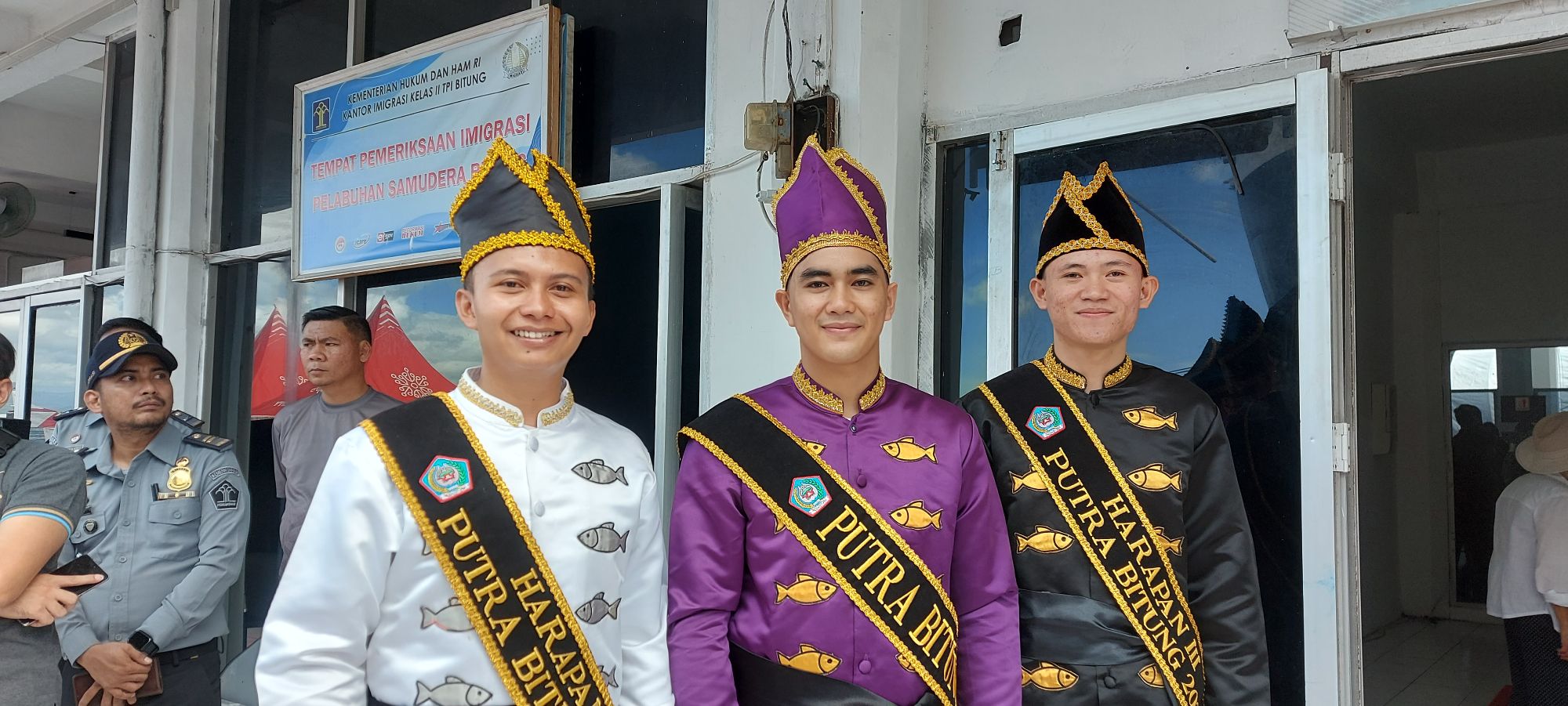 Three young men in traditional Indonesian ceremonial dress with gold-trimmed hats and fish-patterned shirts at immigration office