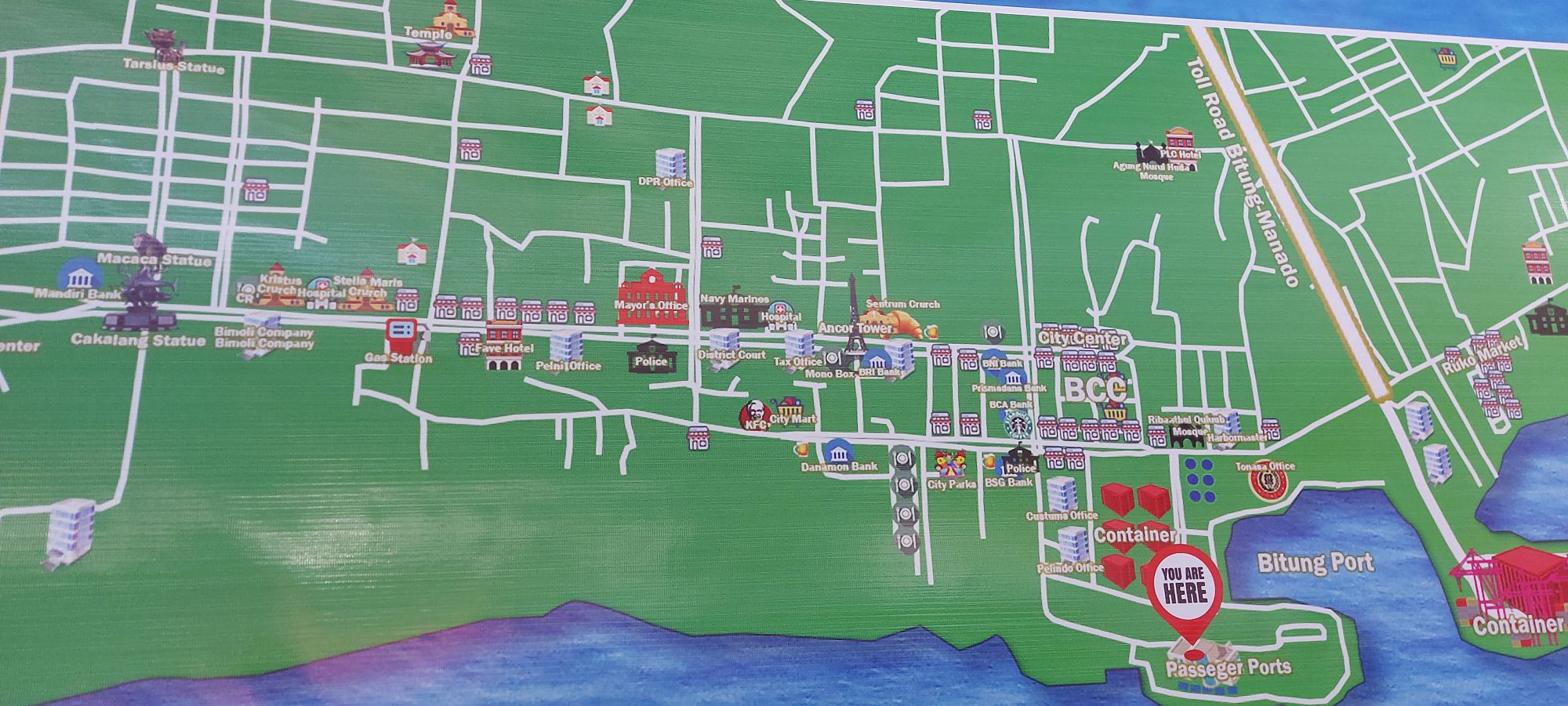 Colorful map of Bitung showing port facilities, city center, and landmarks with 'You Are Here' marker at passenger terminal