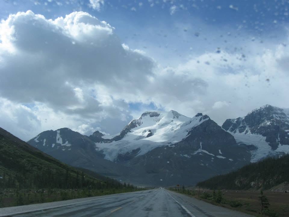 Highway stretching toward snow-capped mountain peaks with coniferous forest on both sides