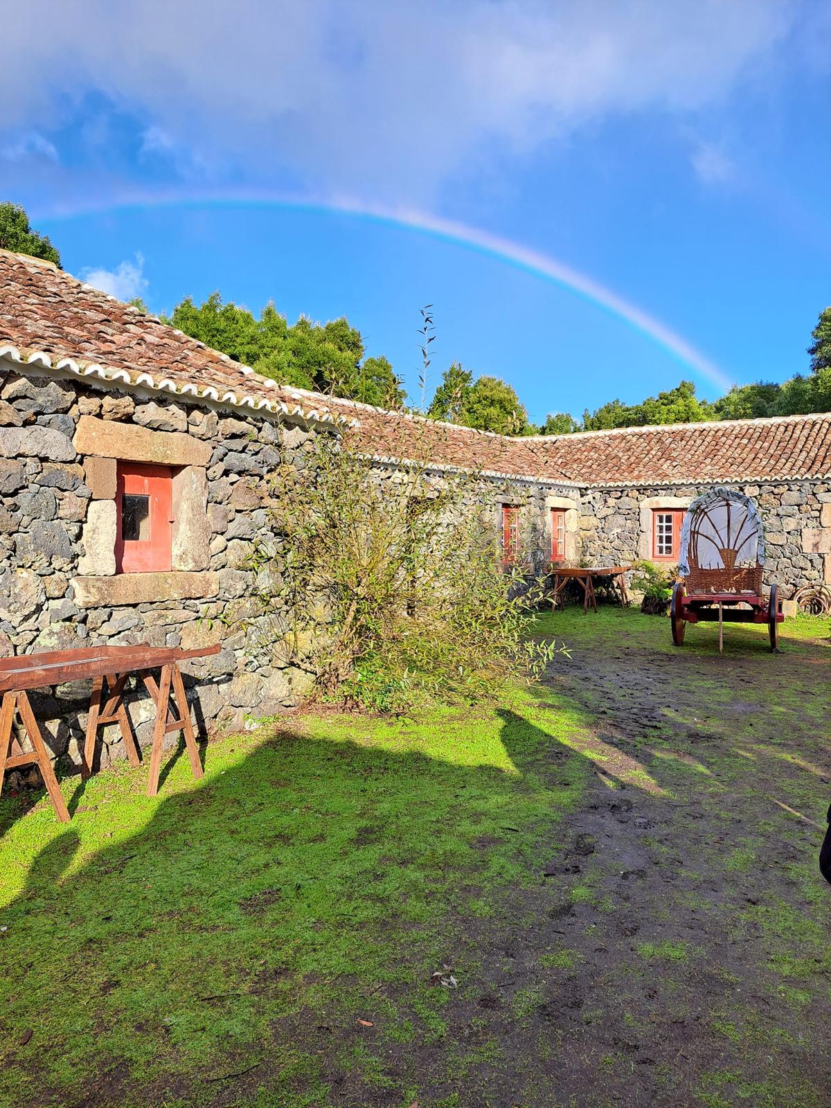Stone farm buildings with red doors and wooden carts in a grassy courtyard under a rainbow in the Azores