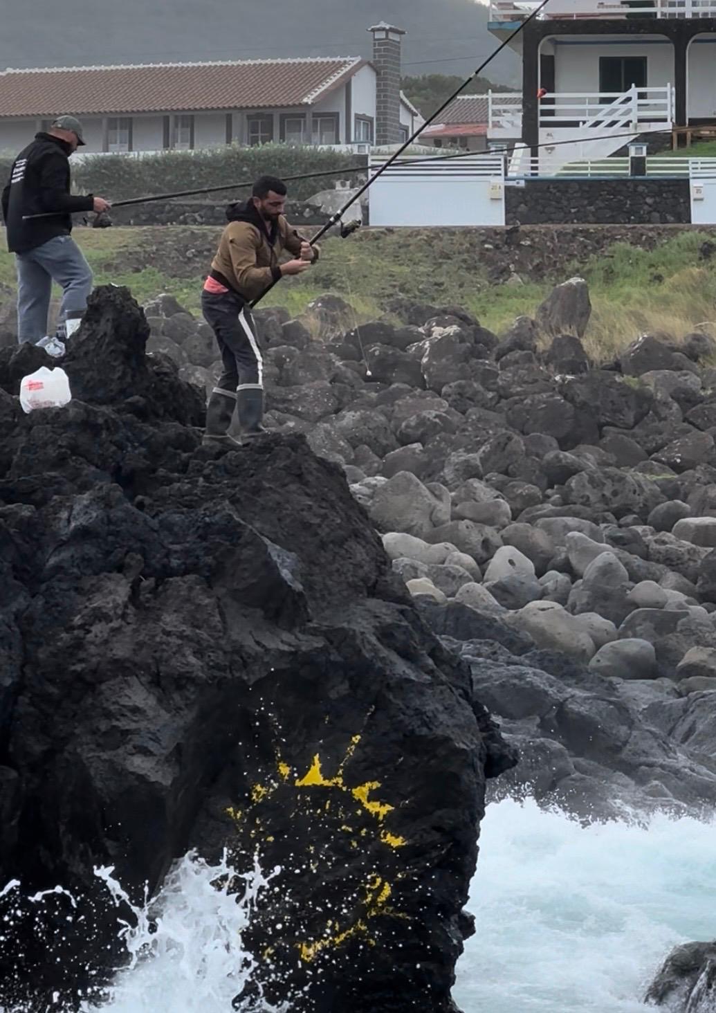 Two people fishing from dark volcanic rocks with white-walled houses visible on the hillside behind them
