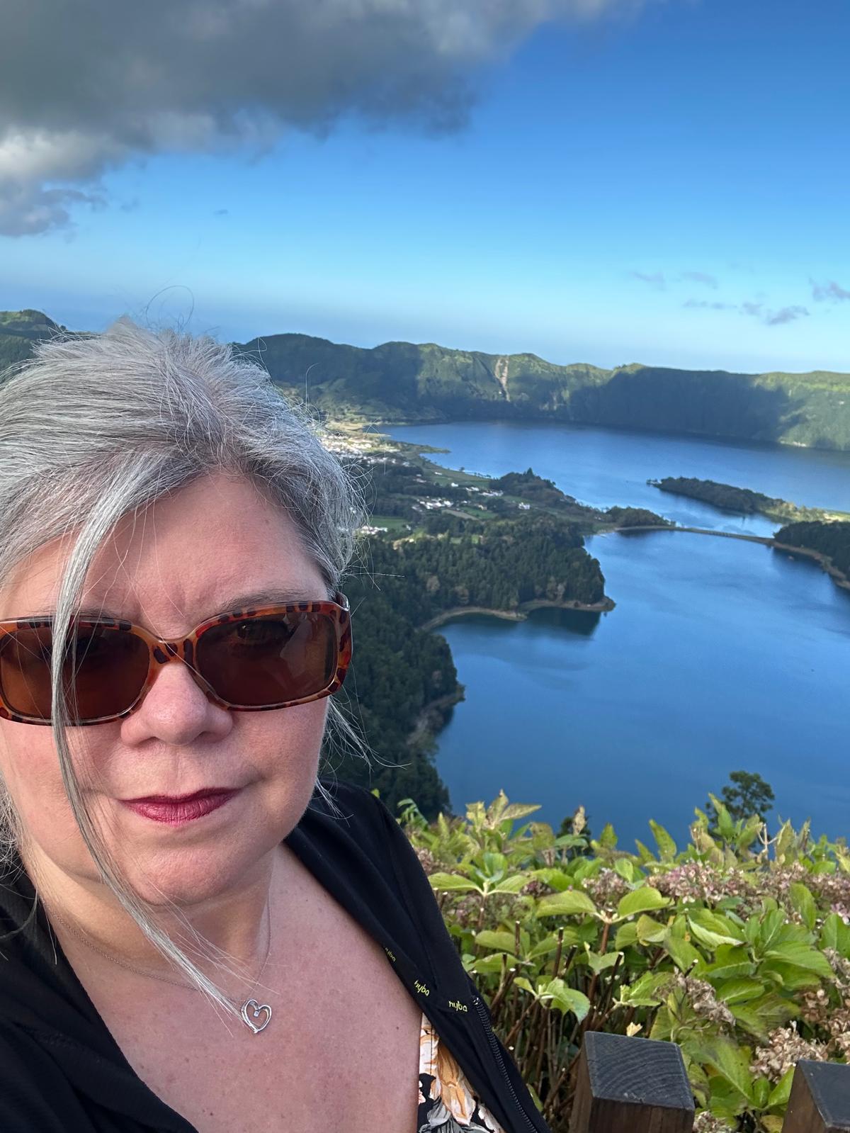 Lesa Nelson smiling at a scenic overlook with a deep blue crater lake and forested volcanic landscape behind her