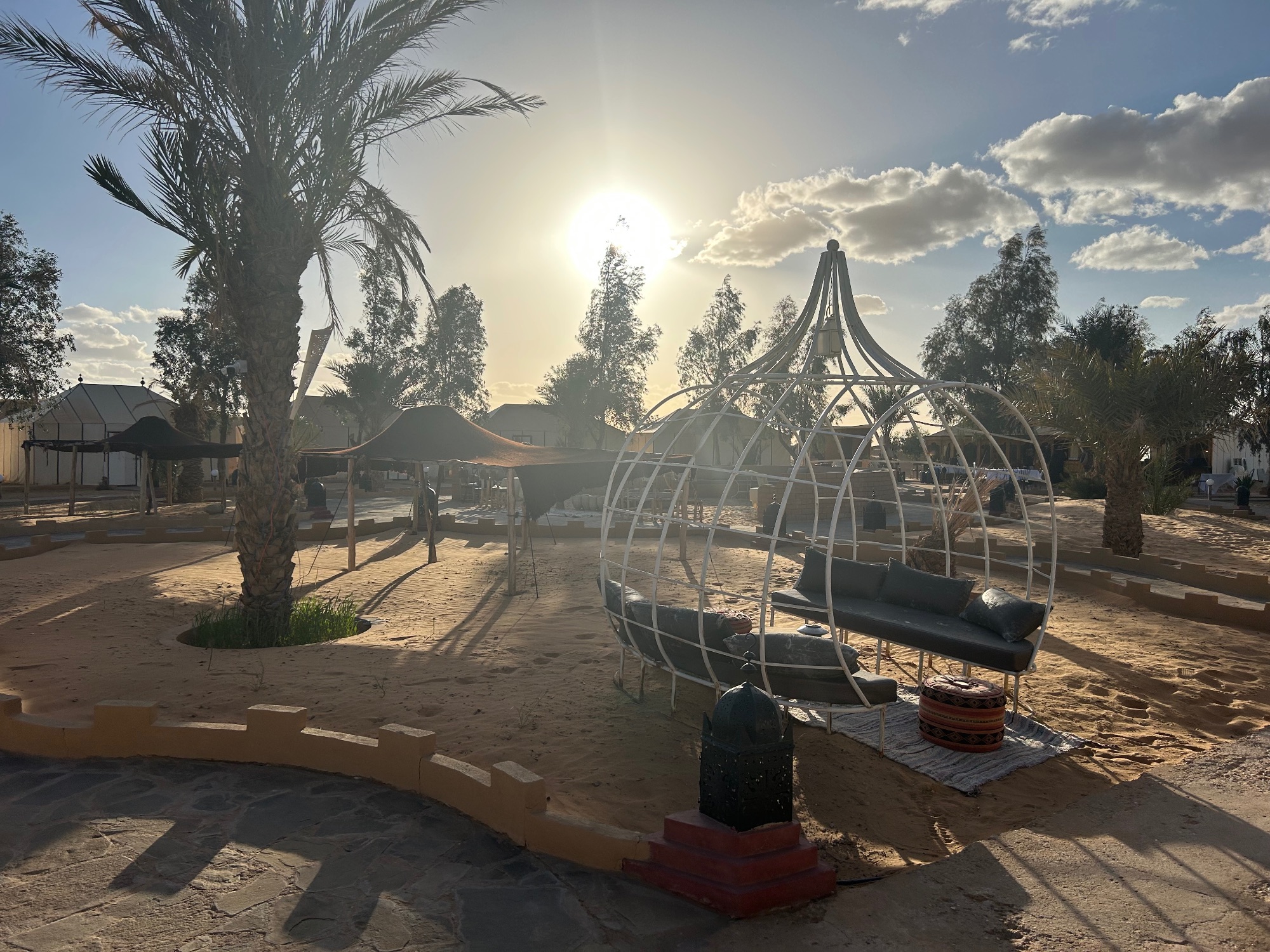 Metal dome-shaped tent structure in a desert camp with palm trees and sandy ground under bright sunlight