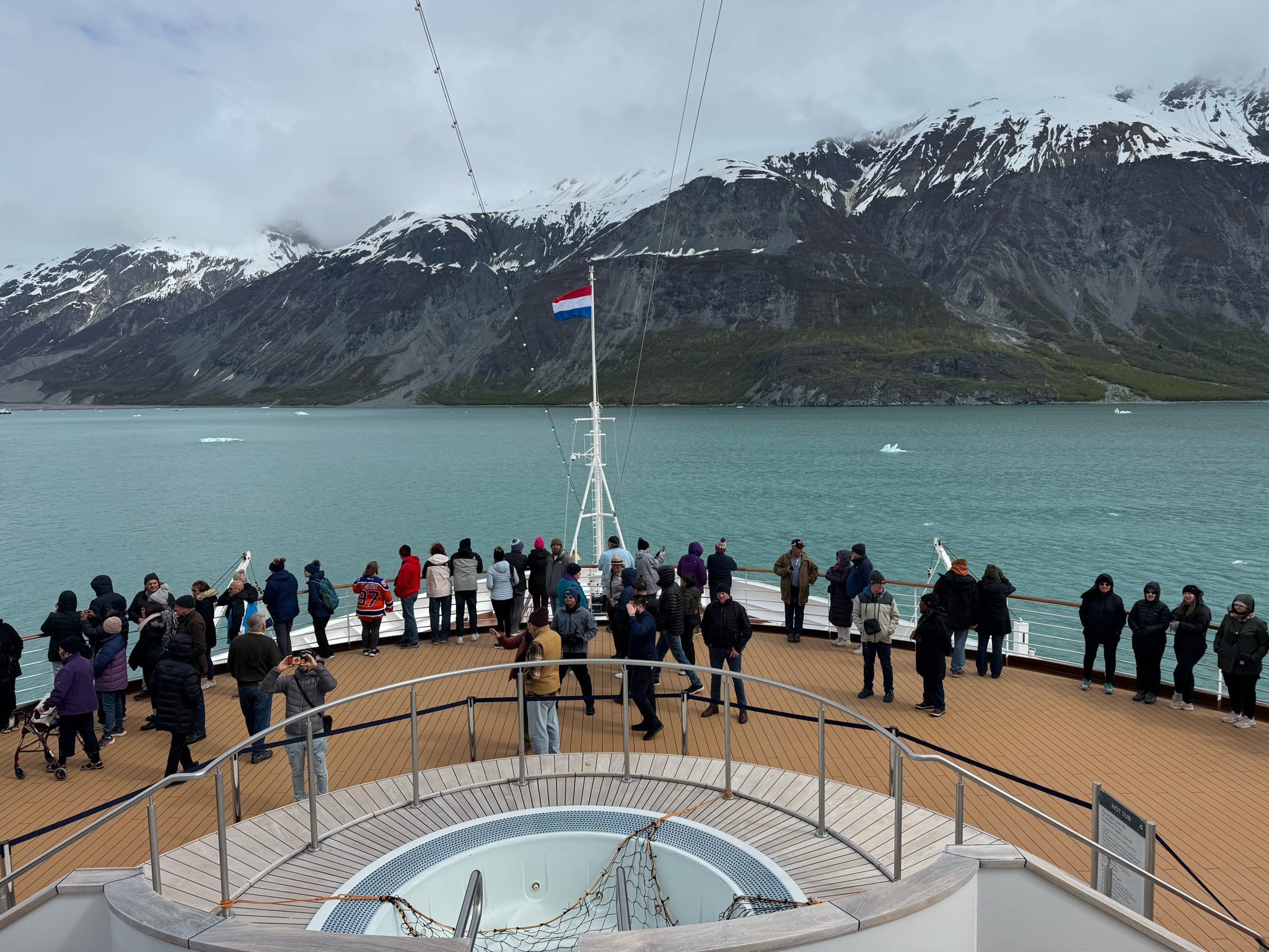 Cruise passengers crowd the bow deck to view dramatic snow-capped Alaskan glacial fjord scenery.