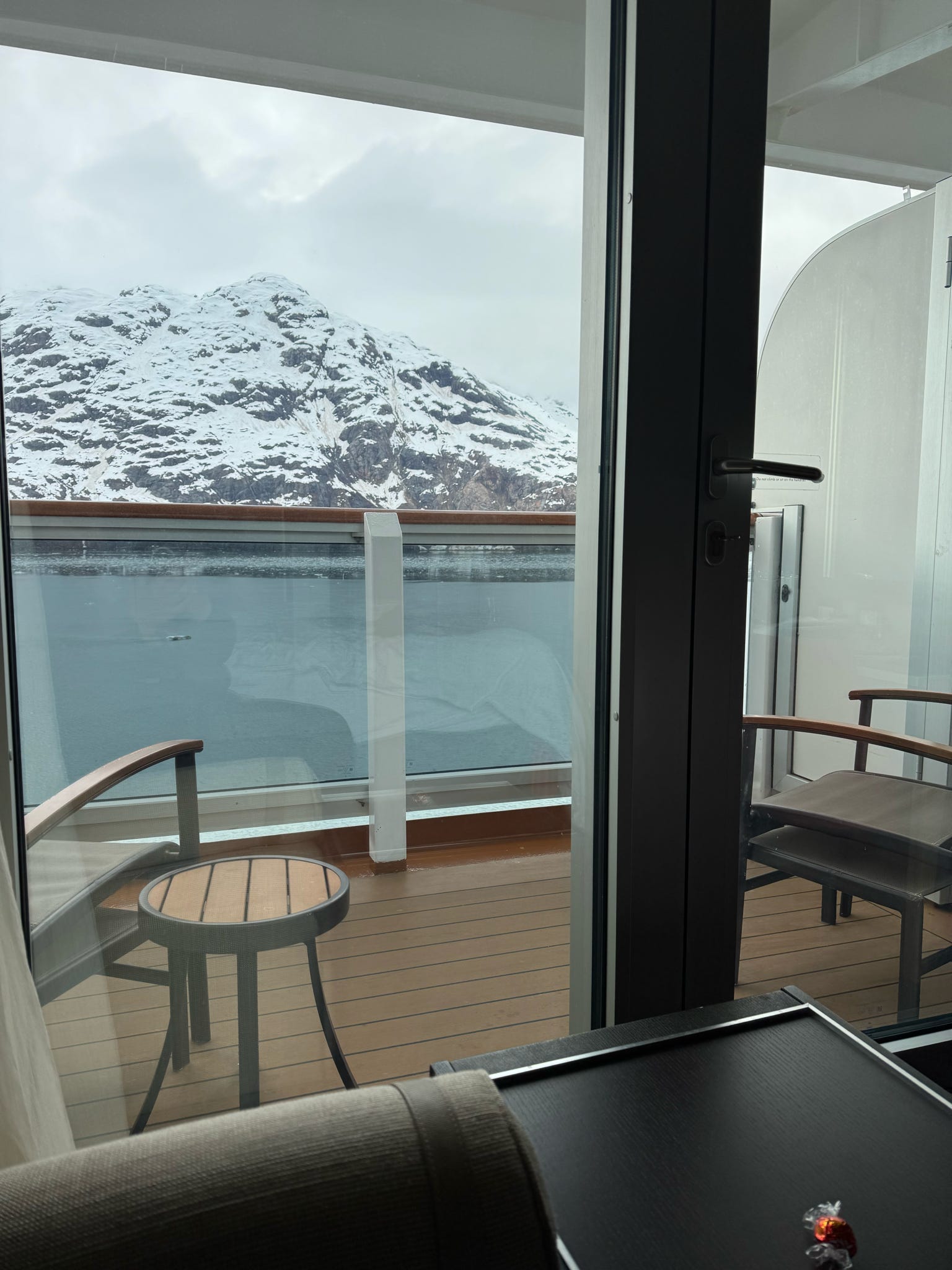 A cruise ship balcony frames a dramatic snow-covered Alaskan mountain rising above icy fjord waters.