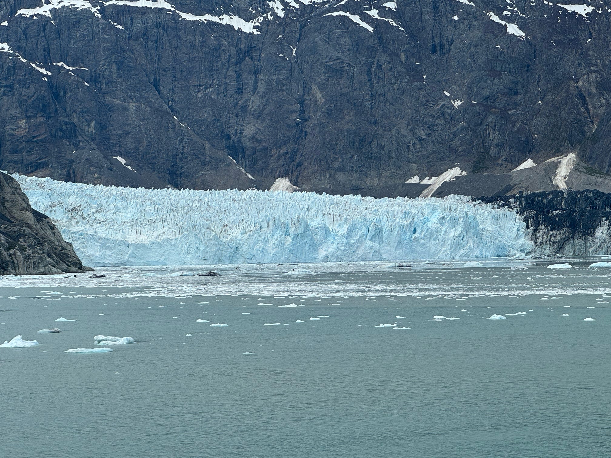 A massive tidewater glacier calves into a frigid Alaskan inlet surrounded by towering dark cliffs.
