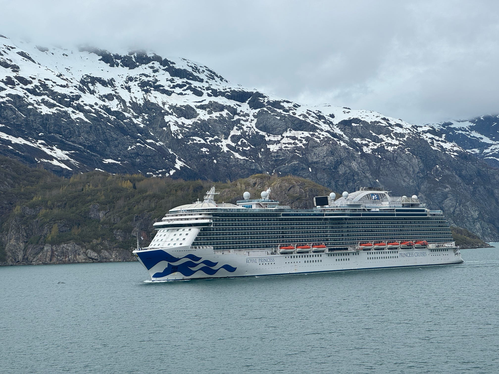 The Royal Princess cruise ship glides through an Alaskan fjord beneath dramatic snow-capped mountains.