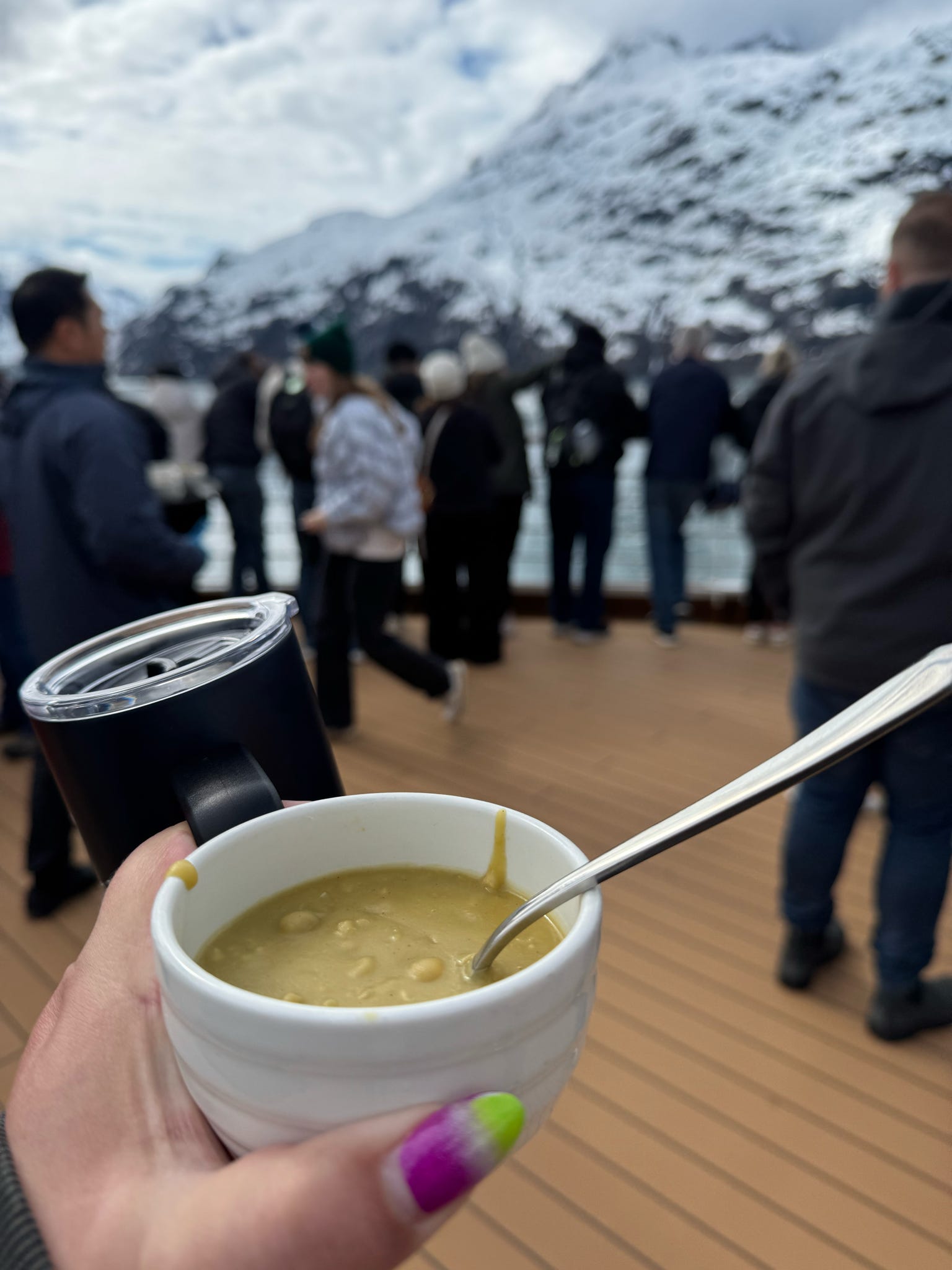 A traveler holds warm soup on a cruise deck with snowy fjord mountains behind.