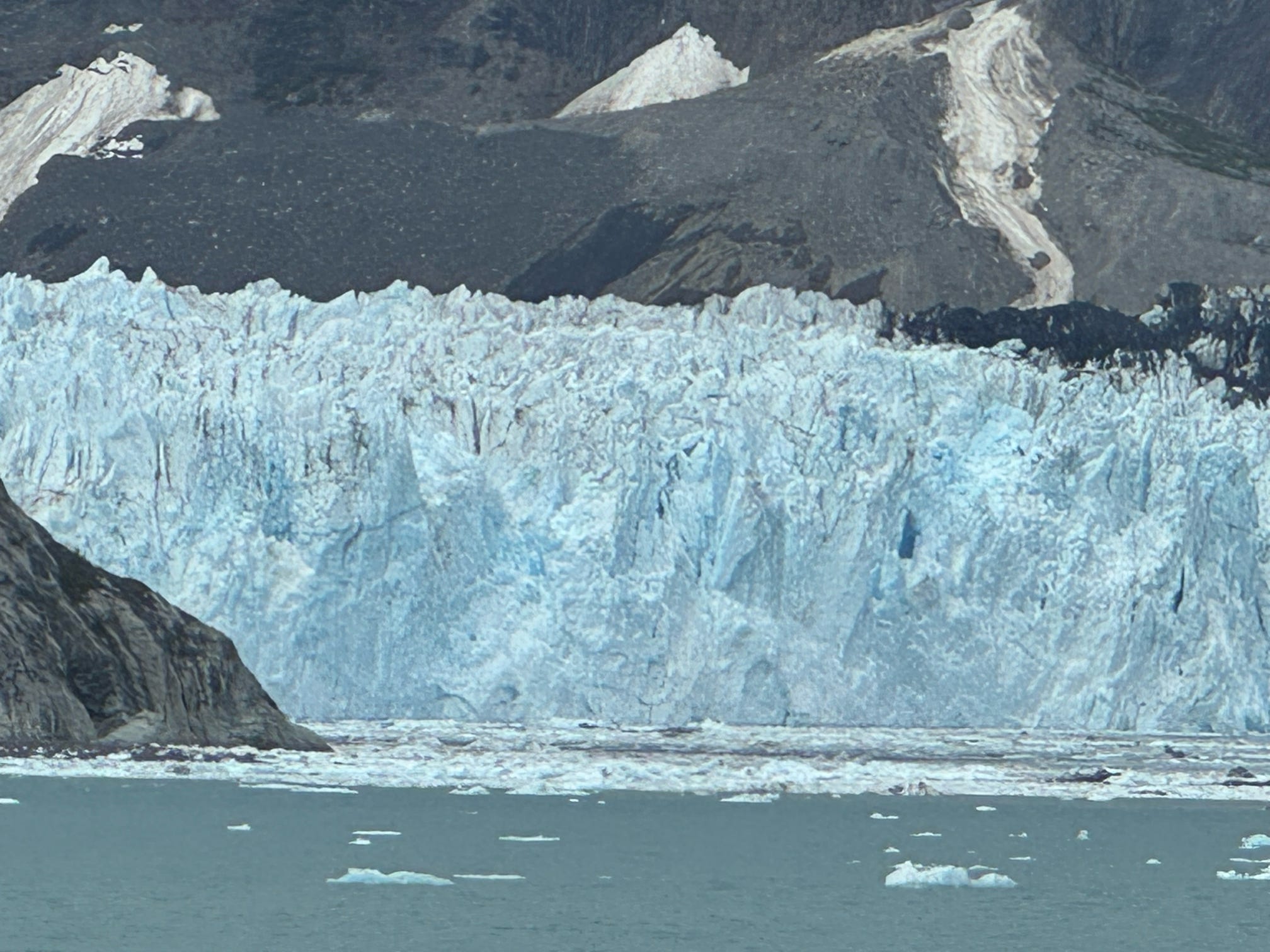 A towering tidewater glacier calving into an icy fjord with rugged mountain peaks behind.