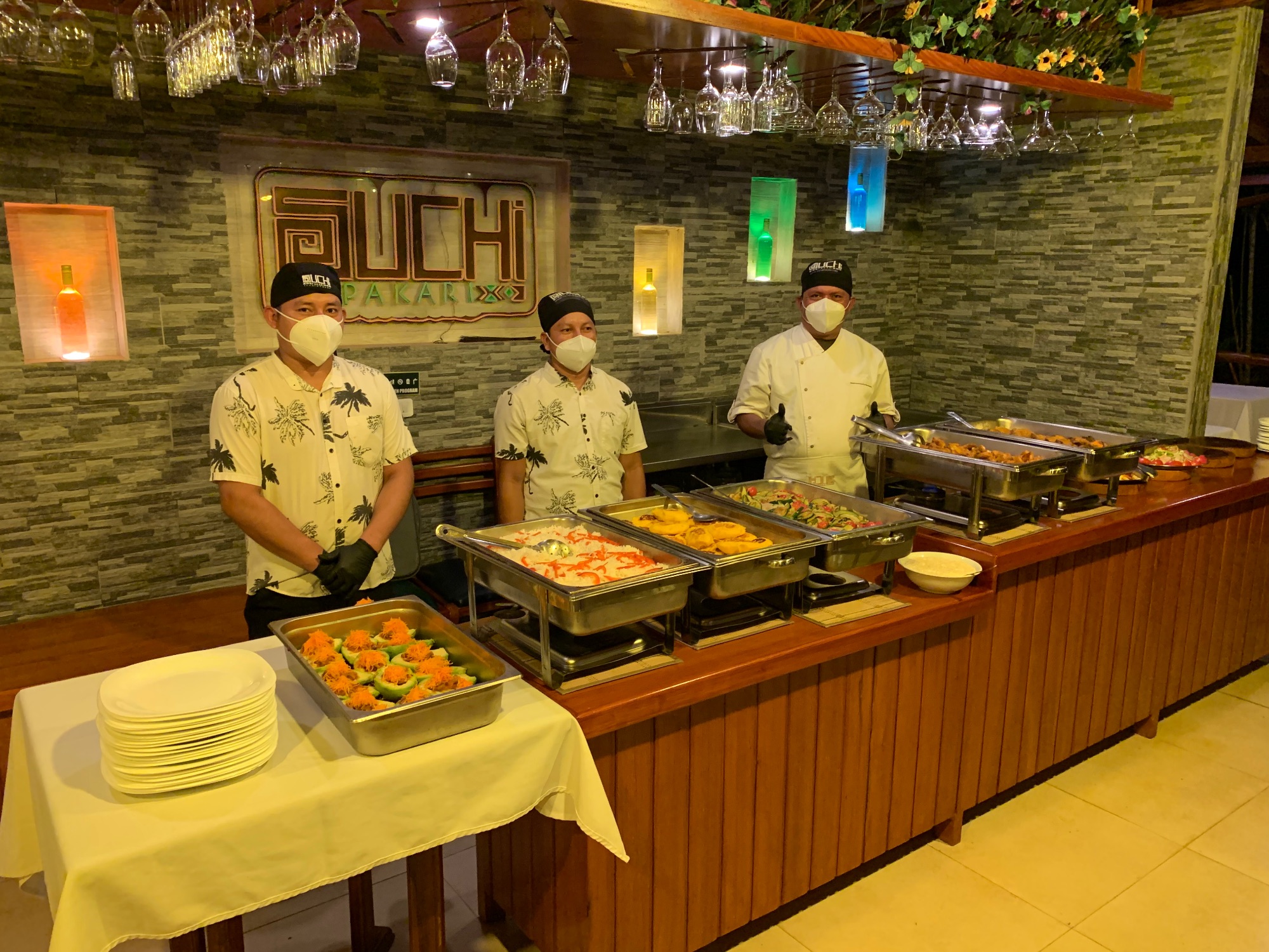 Three staff members in palm-print shirts standing behind buffet counter with food service trays