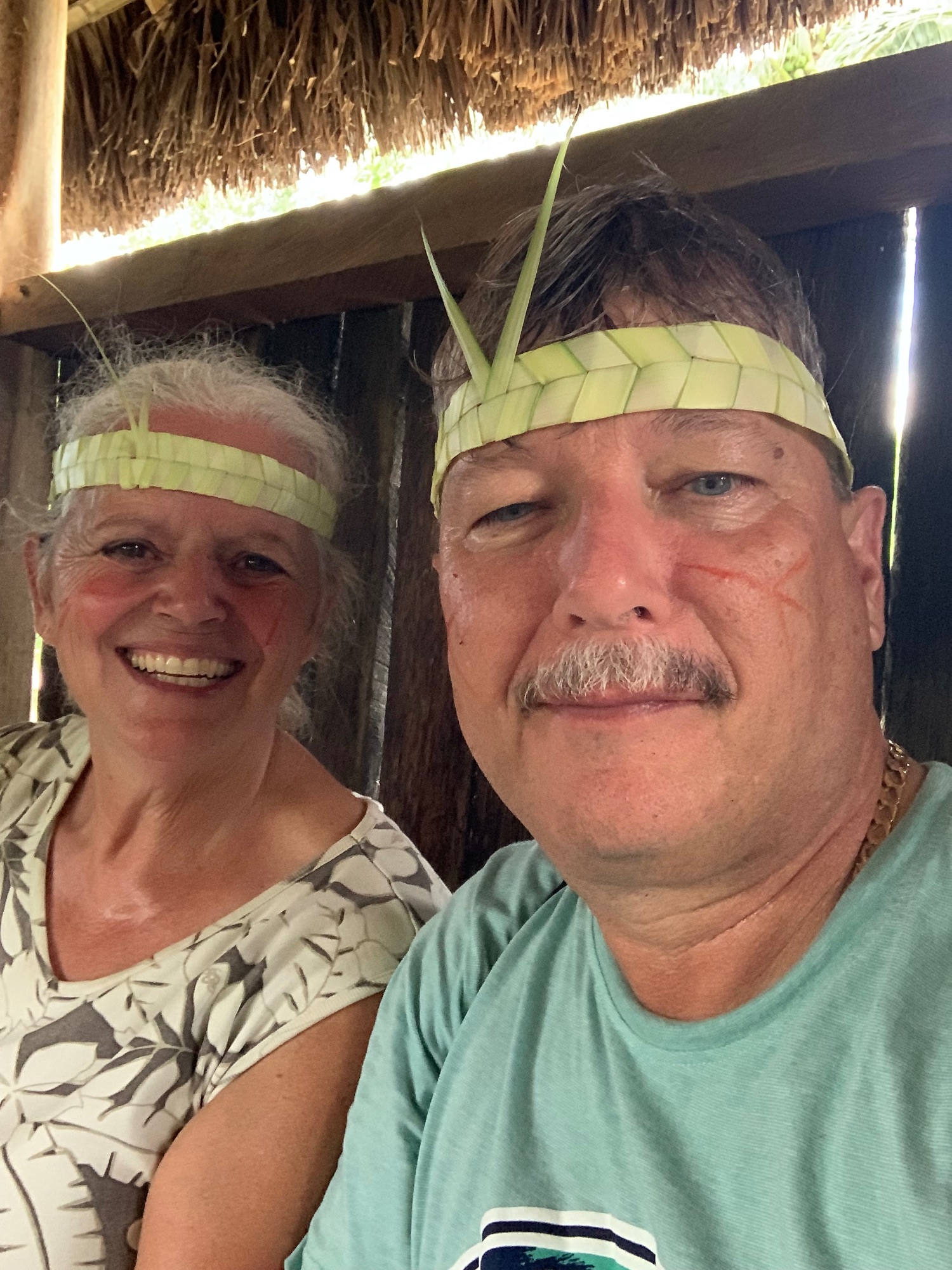 Two people wearing woven palm headbands posing together under thatched roof structure
