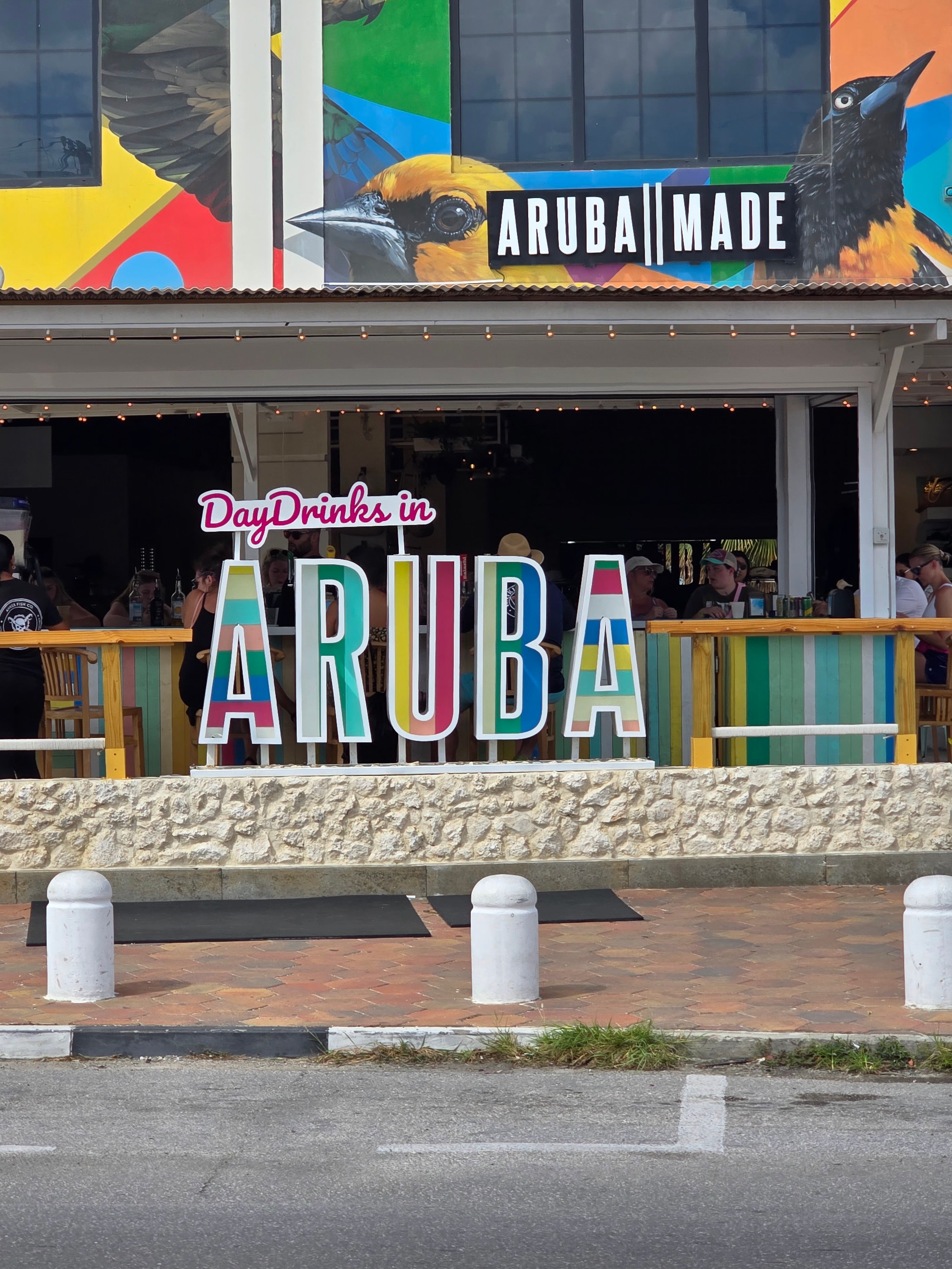 Colorful storefront sign reading 'Aruba Made' with vibrant bird artwork above a beachside bar