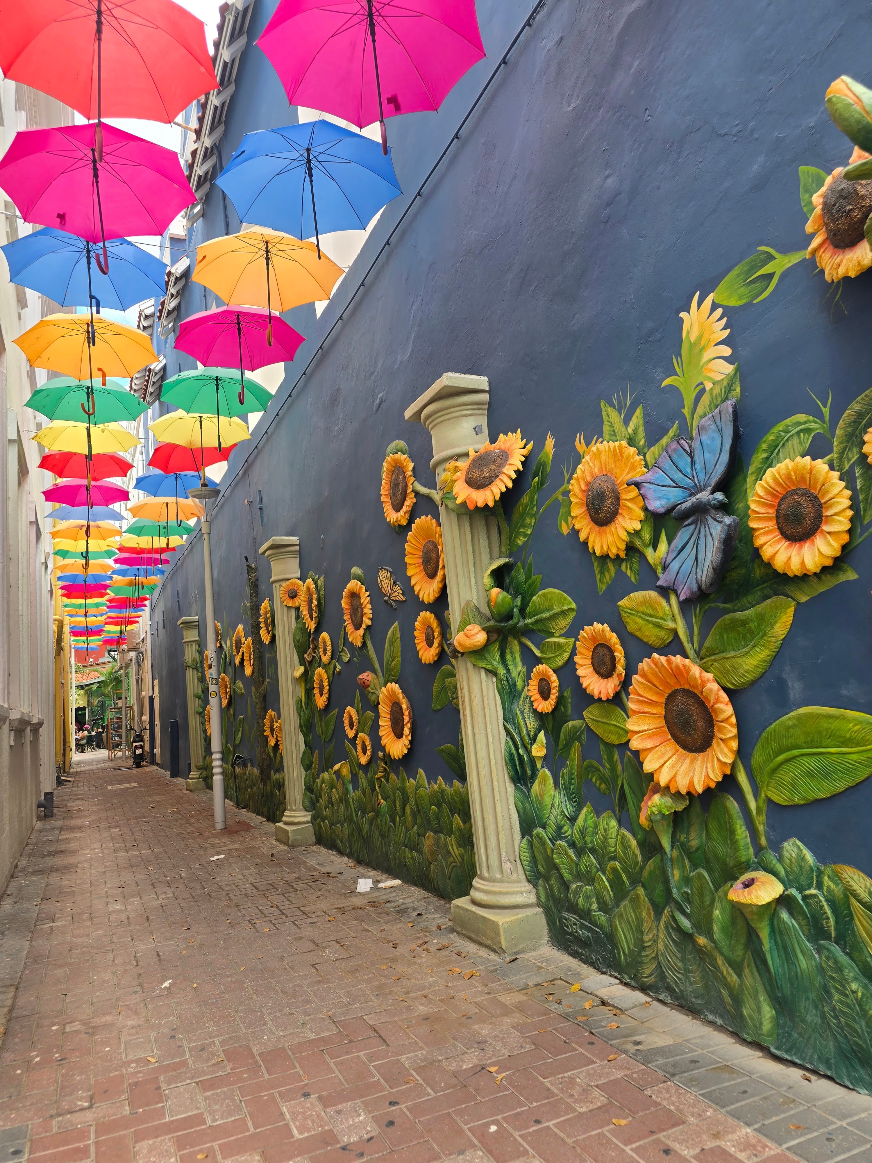 Narrow alley decorated with colorful umbrellas overhead and sunflower mural on dark wall